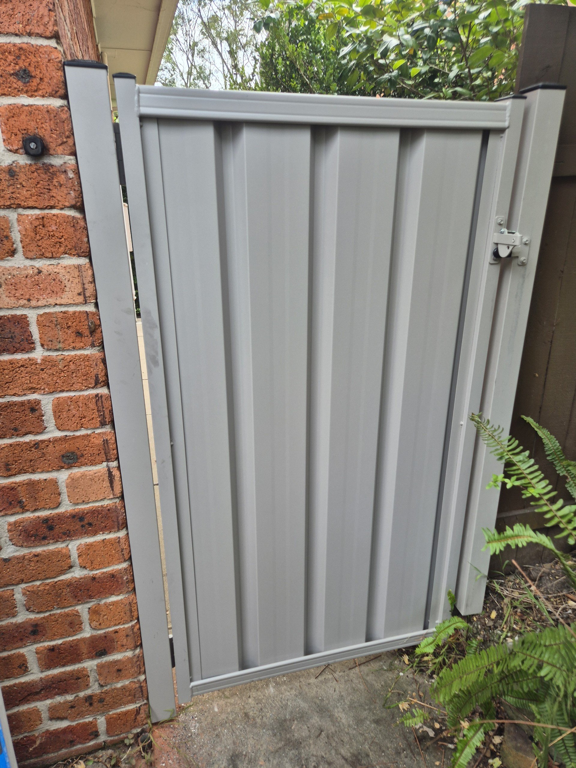 A gray metal gate attached to a brick wall on the left and shrubs on the right, with a latch at the top right corner and some green plants nearby.