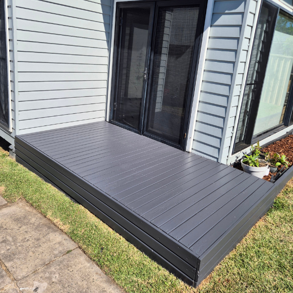Small raised deck with gray composite deck boards outside a sliding glass door next to a garden bed with potted plants.