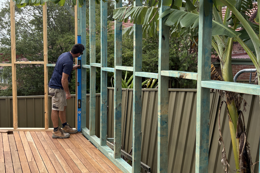 A man measures and levels a wooden frame for a deck under construction outdoors, with a banana tree and a metal fence in the background.
