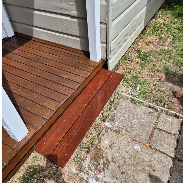 A small wooden step with a darker stained wood plank on a wooden deck near the corner of a house, with concrete paving stones and patches of grass nearby.