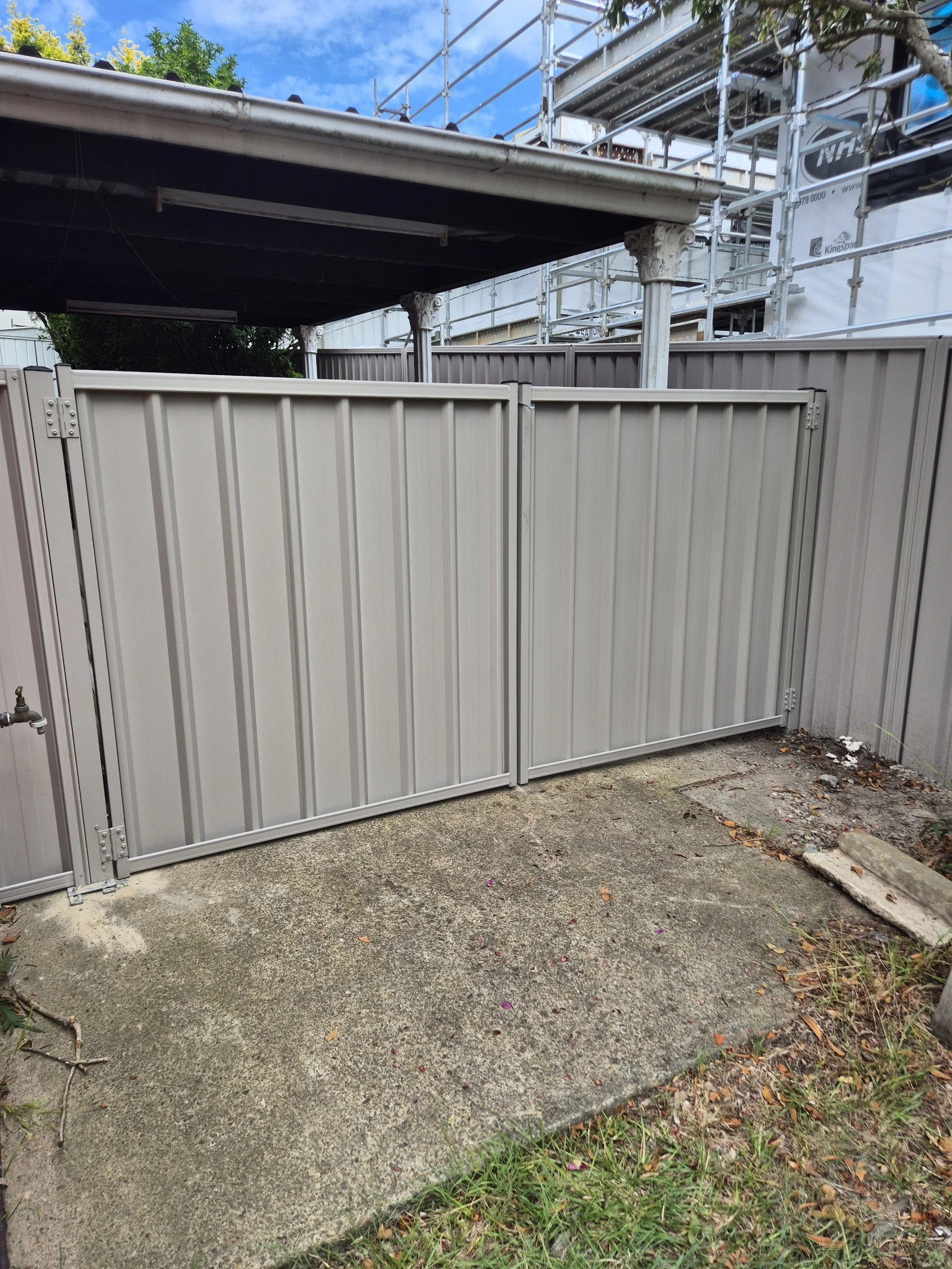A gray metal fence with a gate in a backyard, with potted plants, scaffolding on a building, and a partly cloudy sky visible in the background.