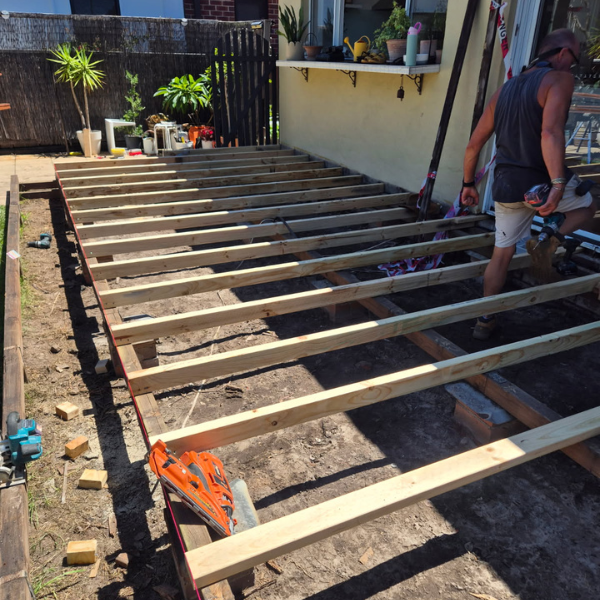 A person working on a wooden deck construction in a backyard, with wooden beams laid out and tools nearby.