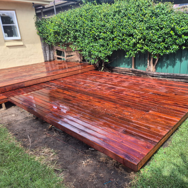 Freshly installed stained wooden deck in backyard with green bushes and a house wall nearby.