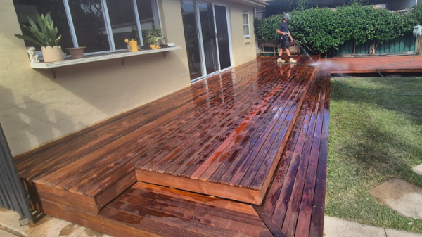 Person power-washing a wooden deck outside a house with large windows and potted plants on the windowsill.