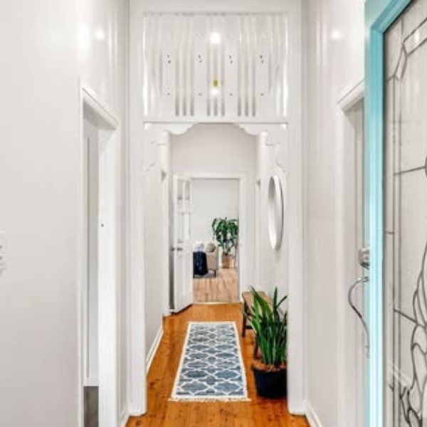View of a narrow hallway with white walls and wooden flooring, leading to a room with a large plant and another smaller plant on the floor, a patterned runner rug, and a doorway in the background.