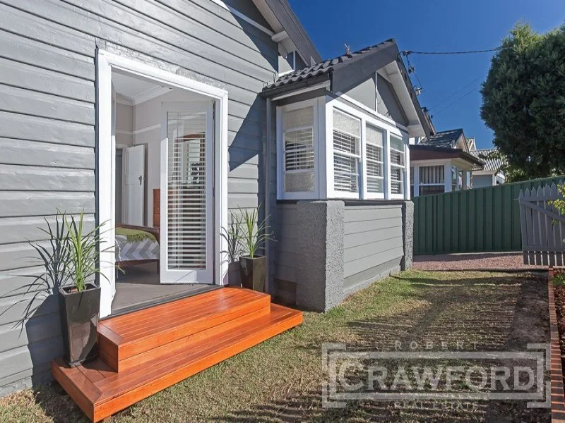 Backyard view of a house with a sliding glass door, wooden steps, potted plants, and a fenced yard, under a clear blue sky.