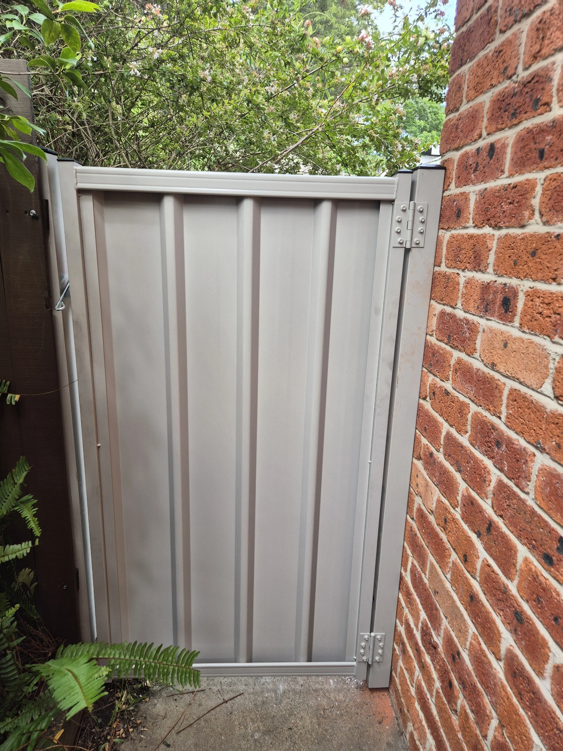 A white metal fence with hinges attached to a brick wall, surrounded by green foliage and plants.