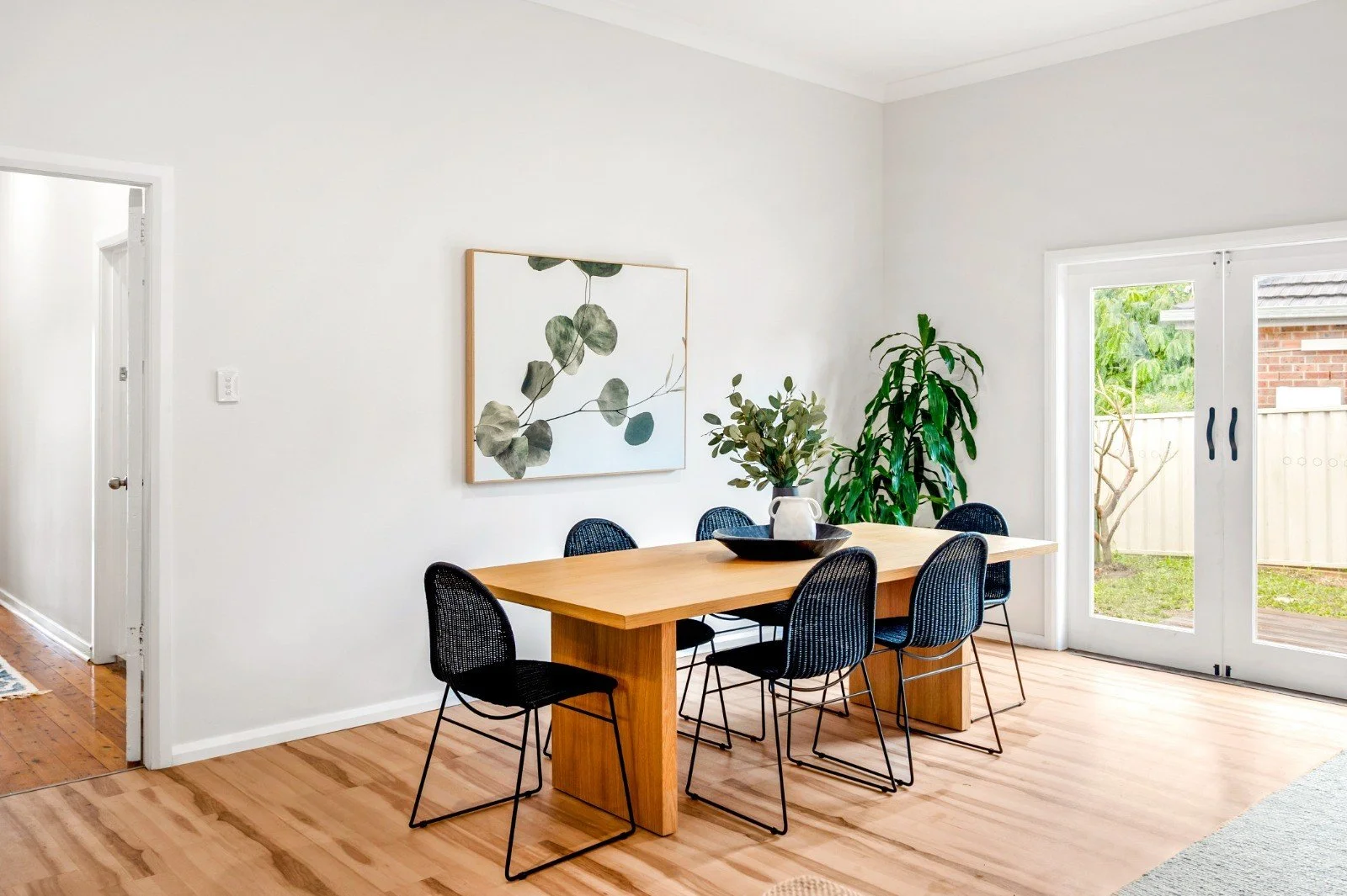 A dining area with a wooden table, six black chairs, a painting of leaves on the wall, and large glass doors leading outside with greenery visible.