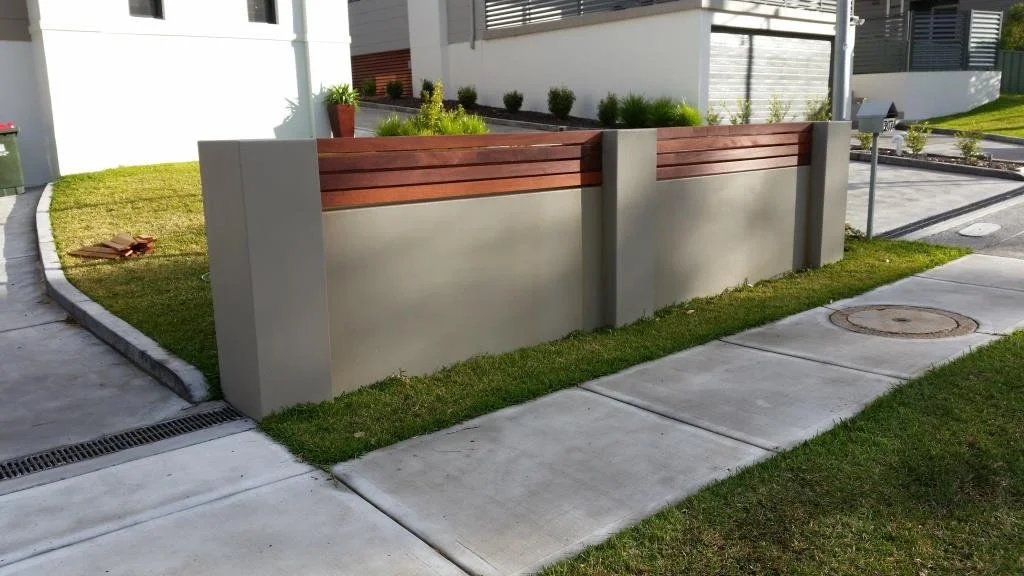 Modern residential outdoor space with a concrete sidewalk, a retaining wall with wooden slats, a small garden with bushes, and a grassy area.