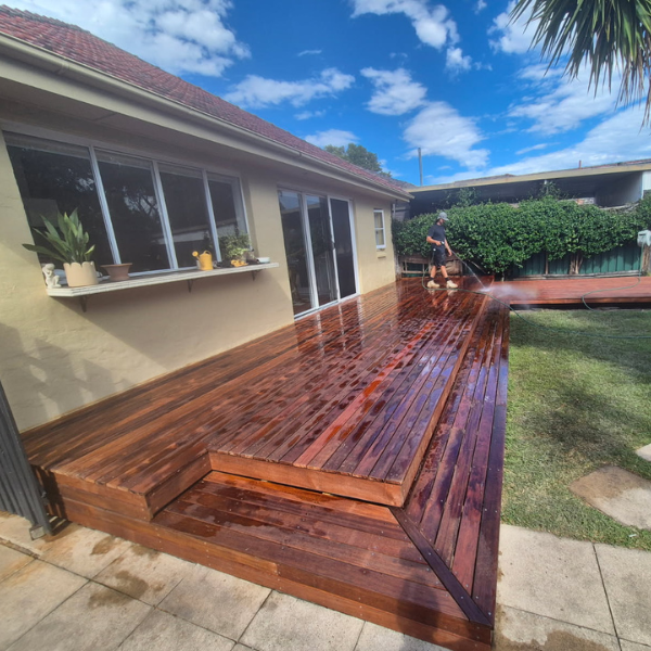 A man power-washes a wooden deck outside a yellow house with large windows and a sliding glass door, under a partly cloudy blue sky.