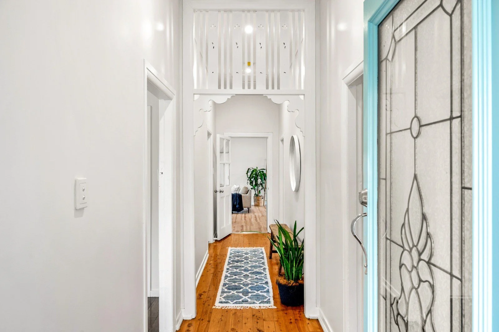 View from a hallway into a living room, with white walls, hardwood floors, a door open to the living room, and a decorative stained glass door on the right. A door at the end of the hallway is partially open, revealing a dining or seating area with a potted plant and a light-colored sofa.