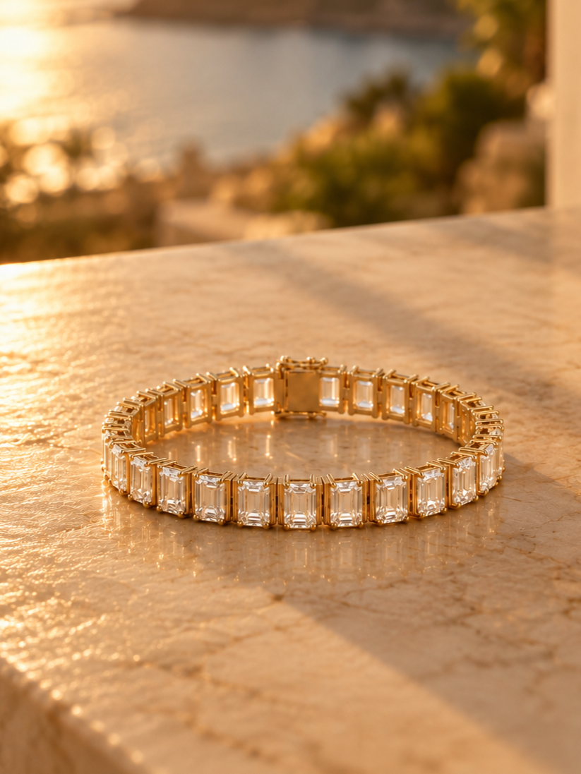 The Emerald Cut gold bracelet with rectangular clear gemstones laid on a textured surface, with a blurred background of water and trees during sunset.