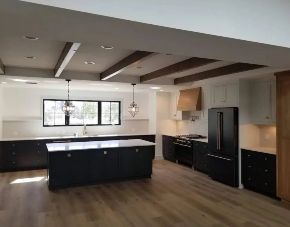 Modern kitchen with black cabinets, a large island, wooden ceiling beams, and pendant lighting over the island. There is a large window above the sink, a black refrigerator, and a stove with a wooden range hood.
