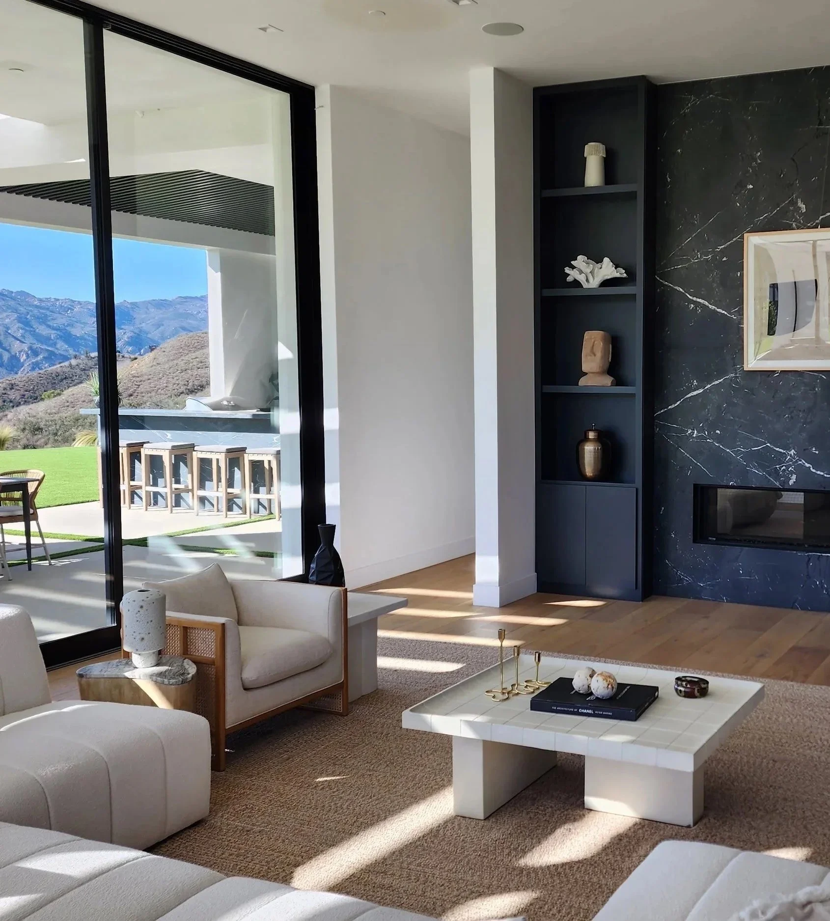 Modern living room with sliding glass doors opening to scenic mountain view outdoor patio with barstools. Interior features a black built-in shelf with decorative items, a white marble coffee table with books and ornaments, beige seating, and a beige rug. Sunlight illuminates the room, highlighting the minimalist decor.