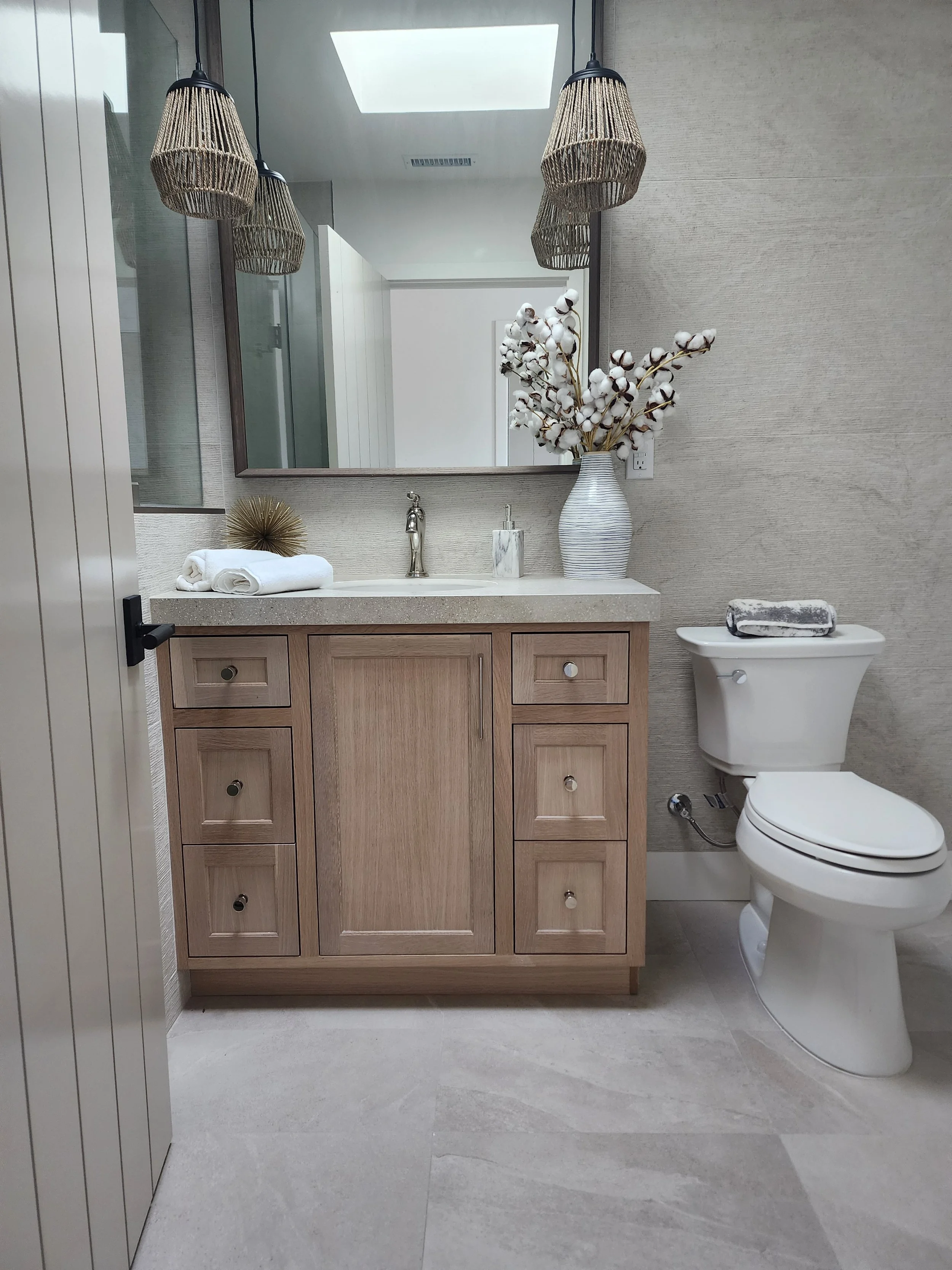 Bathroom with a wooden vanity, a large mirror, cotton flowers in a striped vase, and a toilet with a folded towel on top. Woven pendant lights hang from the ceiling, and skylight and ventilation are visible.