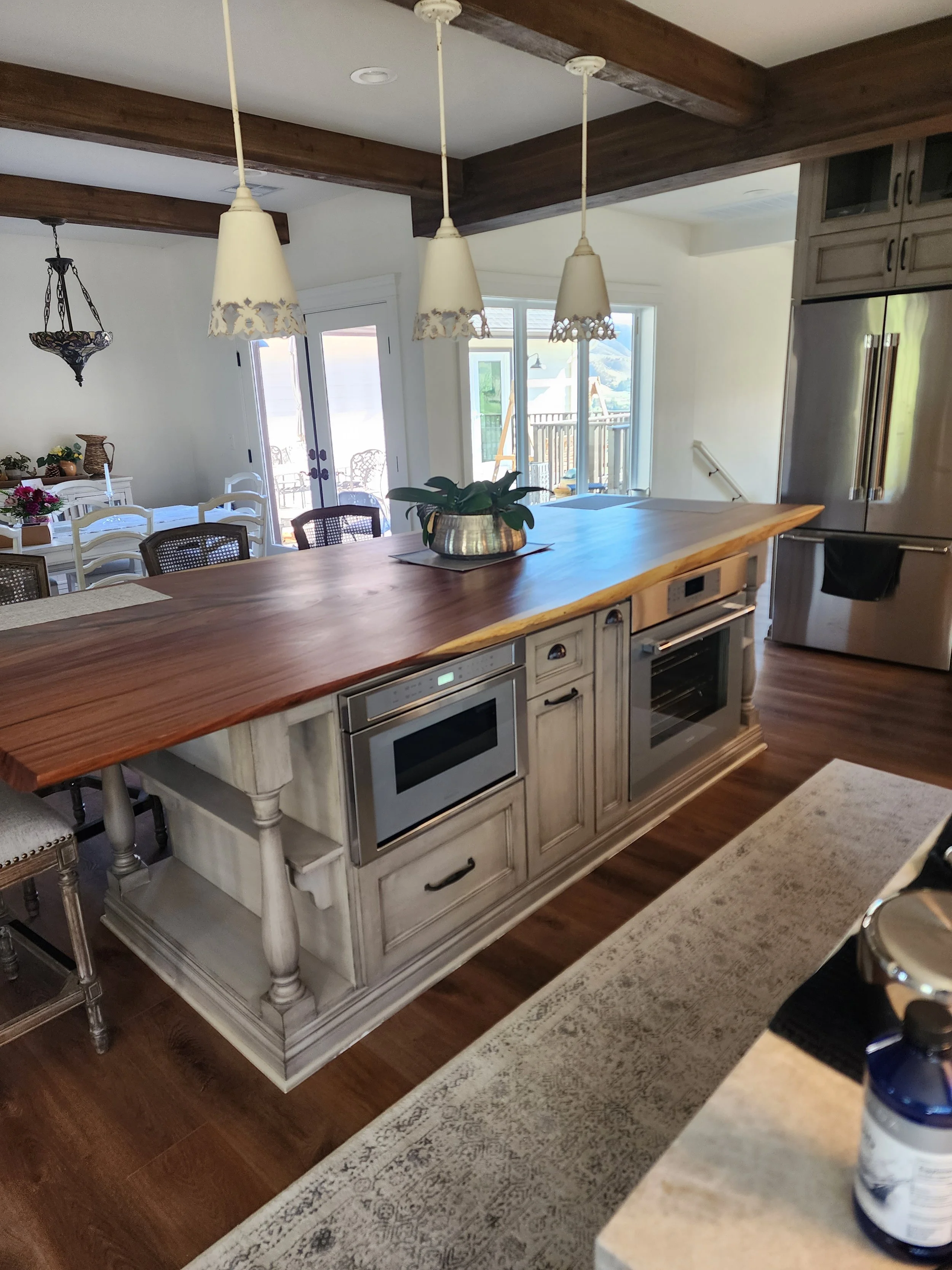 A kitchen with a wooden countertop island, white cabinets, stainless steel appliances, a plant on the counter, and pendant lights hanging from a beam. In the background, a dining table with chairs and a sliding glass door leading outside.