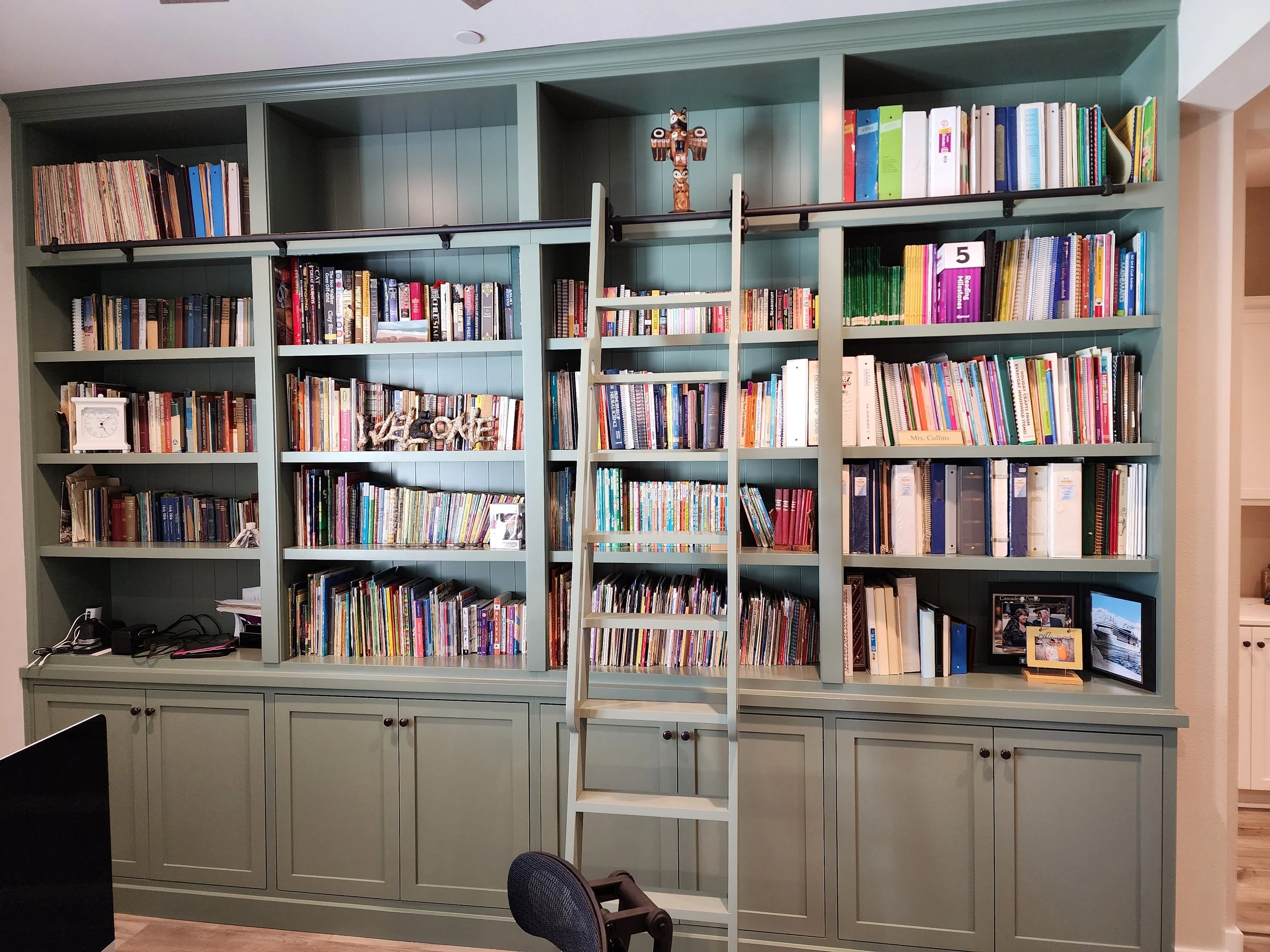 A large bookshelf painted in teal, filled with books and decorations, is situated in a room with wood flooring. There is a small ladder in front of the shelves, a clock, and framed photos on the lower part of the bookshelf, with chairs and a laptop in the foreground.