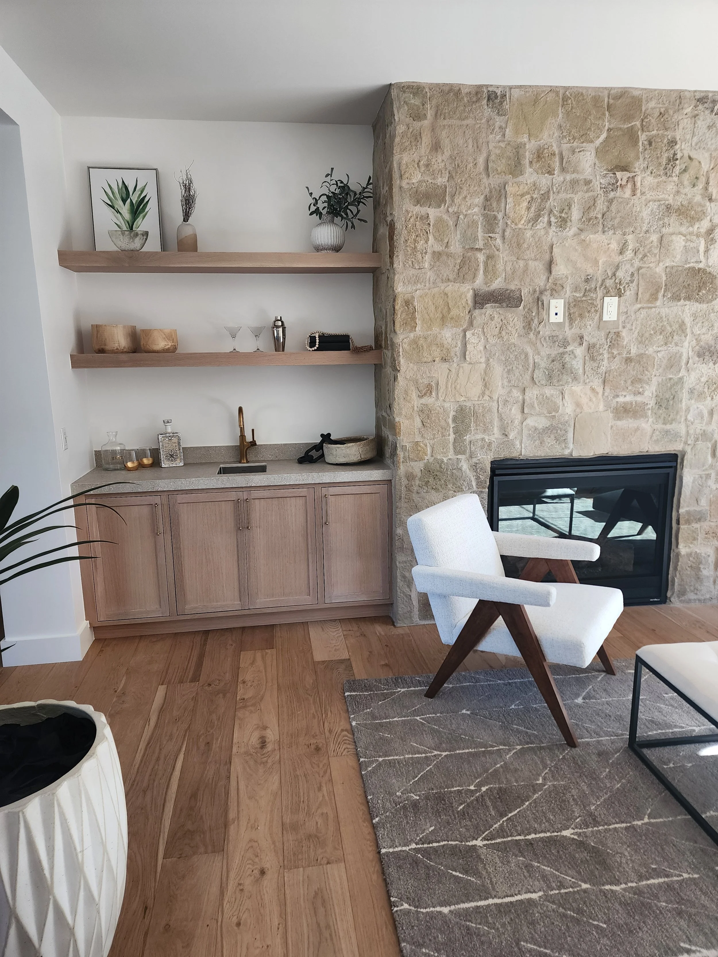 A cozy living room corner with a stone fireplace, a white armchair with wooden legs, a wooden cabinet with a countertop, and two wooden wall shelves decorated with plants, artwork, and decorative objects.