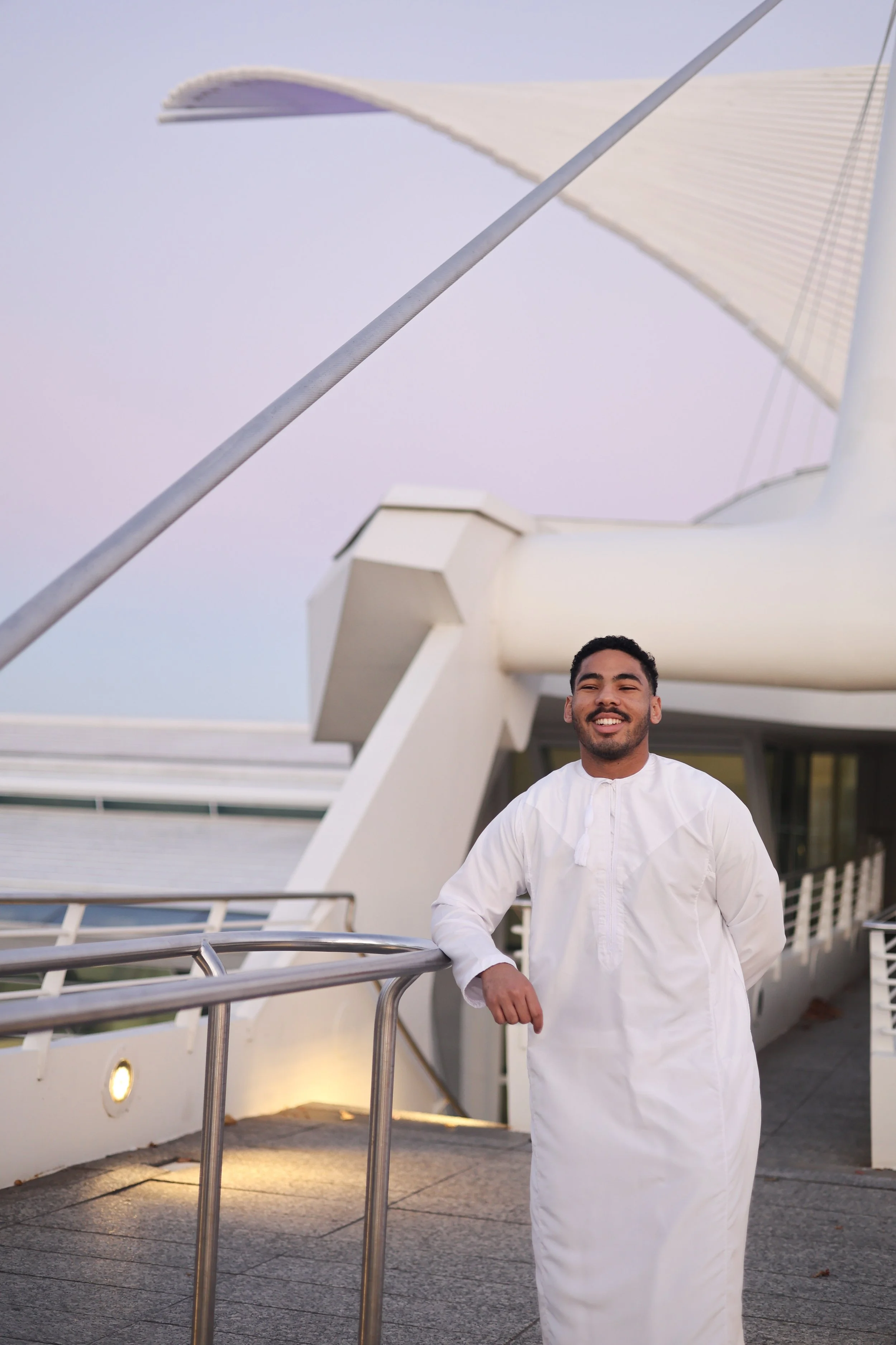 A man wearing a white traditional garment standing outside on a balcony, smiling with a modern building with an architectural structure in the background during dusk.