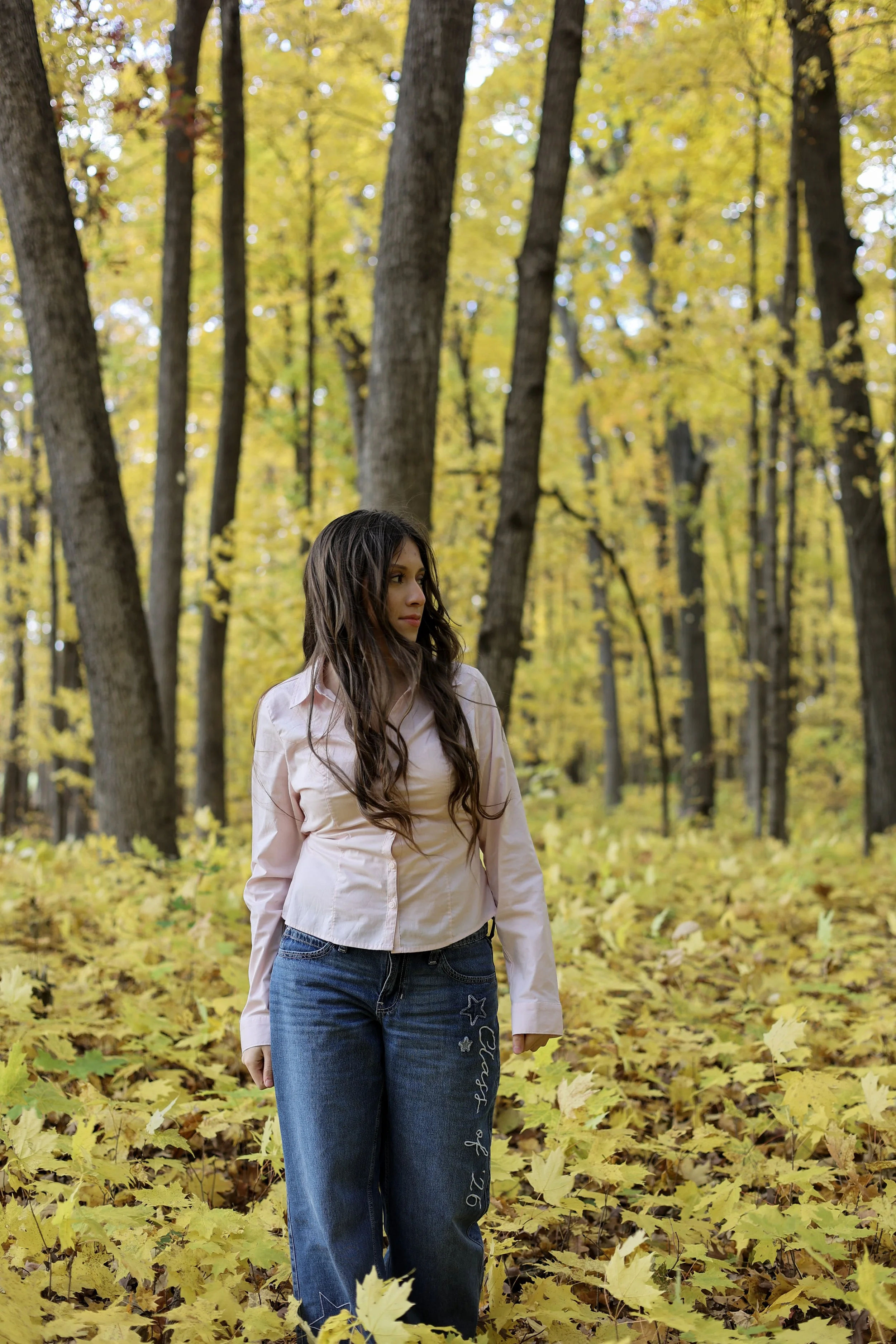 A young woman standing outdoors in a forest with yellow fall leaves on the ground and trees with yellow leaves in the background.
