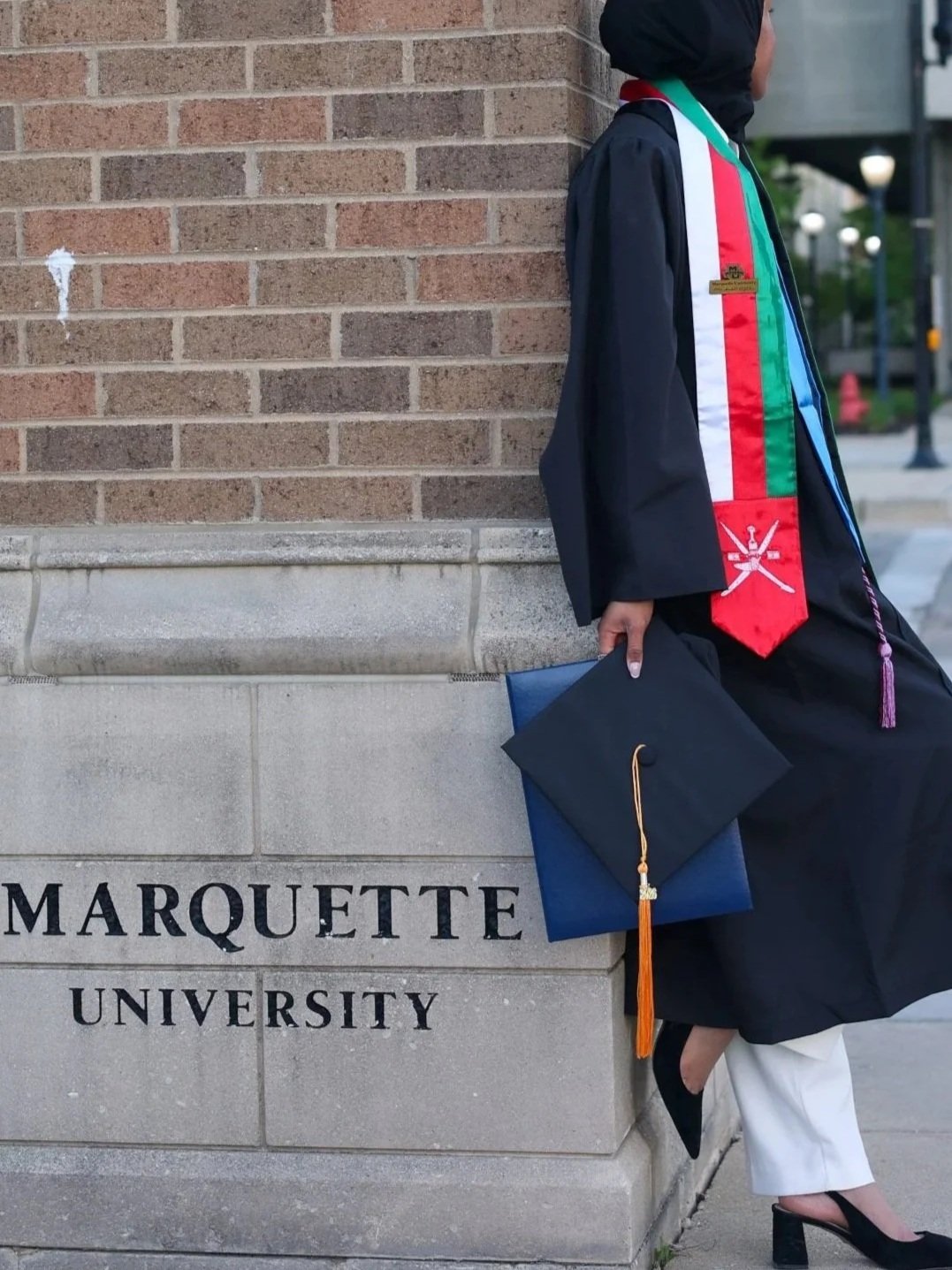 A graduate in a cap and gown standing with their back to a brick wall at Marquette University, holding a rolled diploma, wearing a colorful stole, and wearing black high heels and white pants.