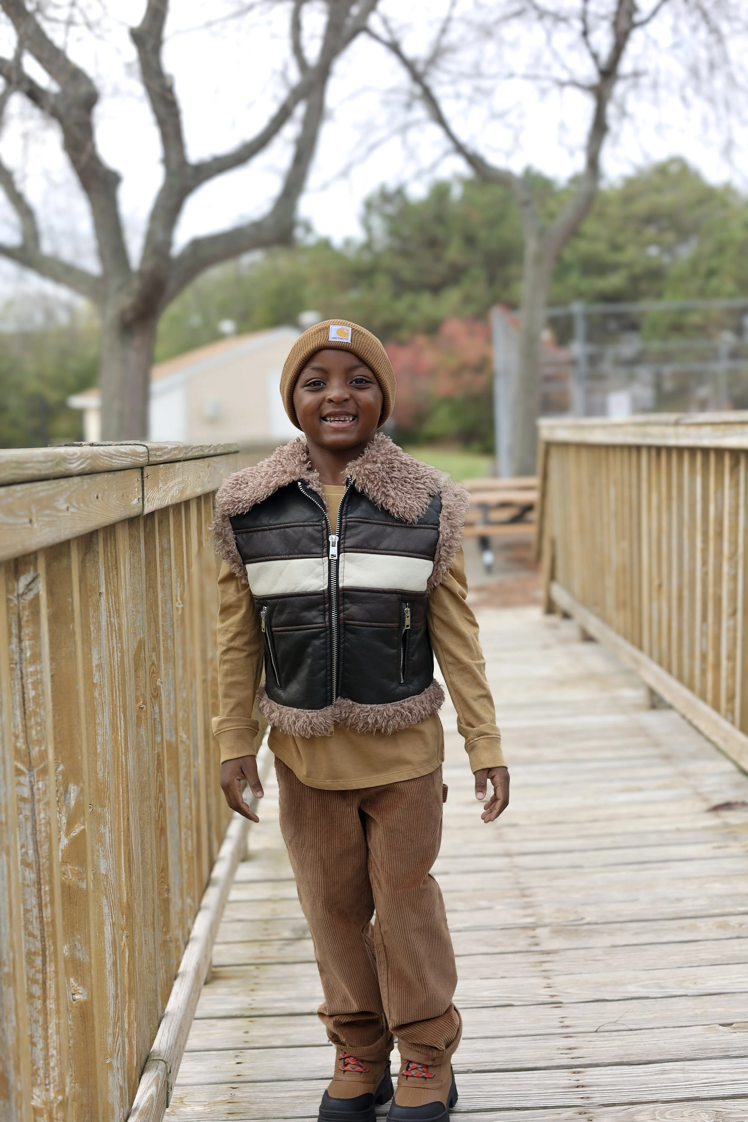 A young boy with a big smile walking on a wooden bridge outdoors during autumn, wearing a brown beanie, a black and beige jacket with furry lining, brown pants, and brown boots.