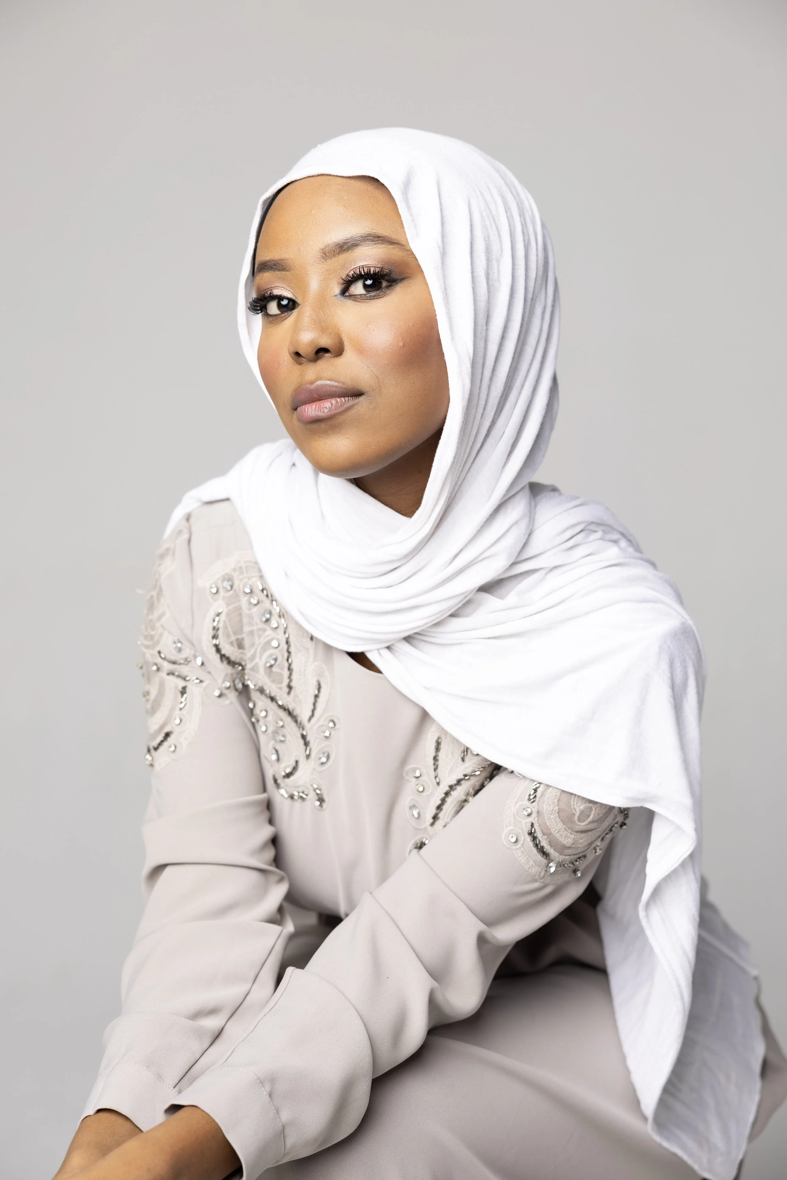 A woman wearing a white hijab and a white top with embroidered details, sitting against a plain gray background.