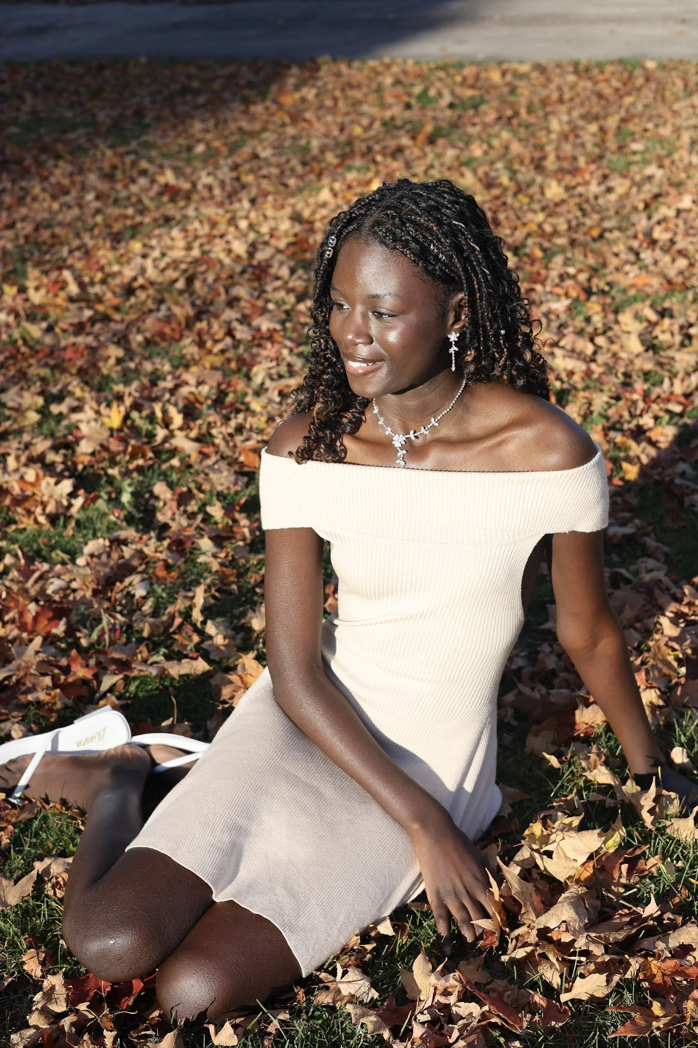 A woman sitting on the fallen autumn leaves, wearing an off-the-shoulder beige dress, with curly black hair styled in braids, accessorized with jewelry, outdoors in a park during daytime.