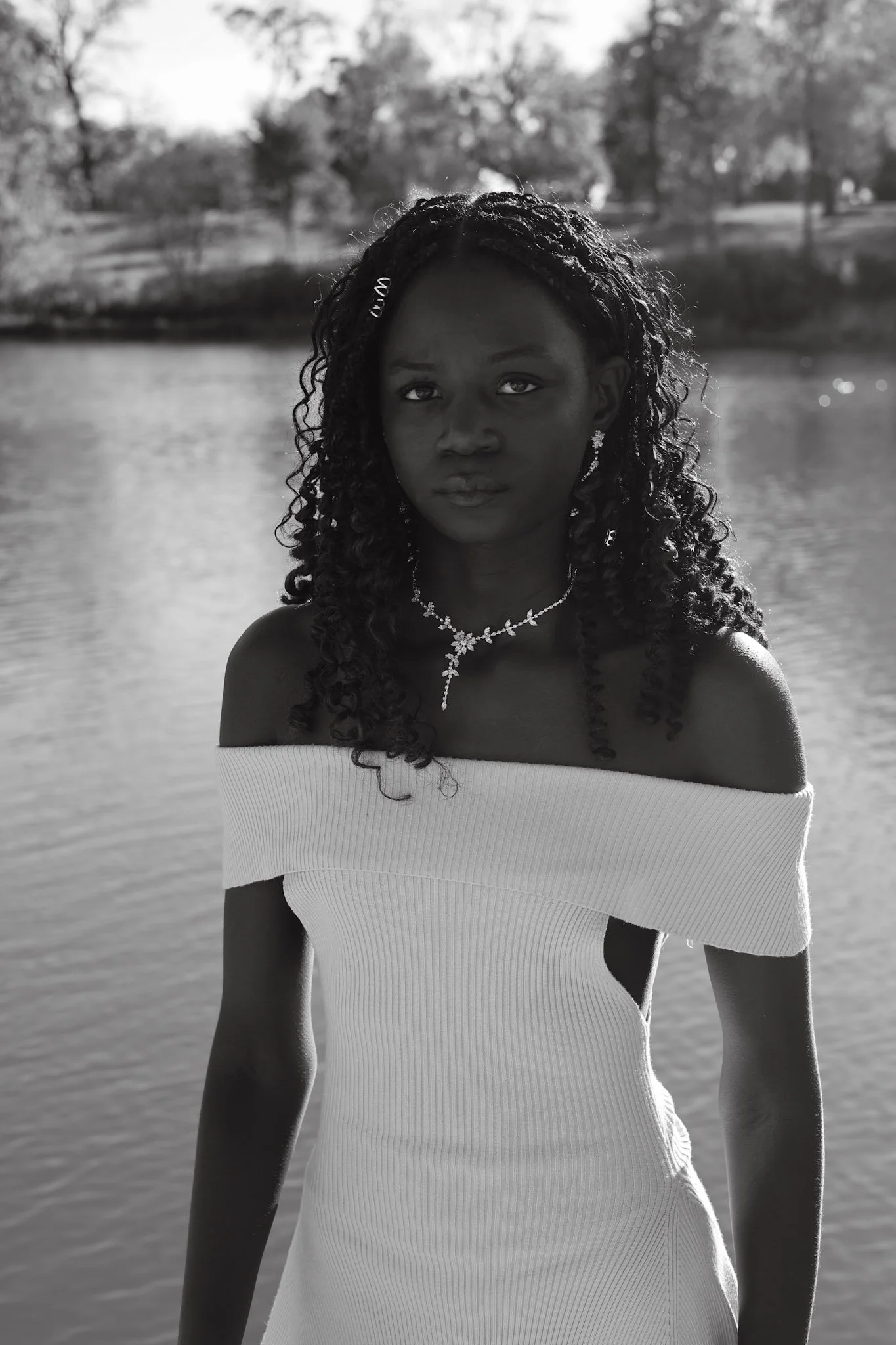 A woman with curly hair wearing a white off-shoulder dress and jewelry stands by a body of water, with trees and a park in the background, in black and white.