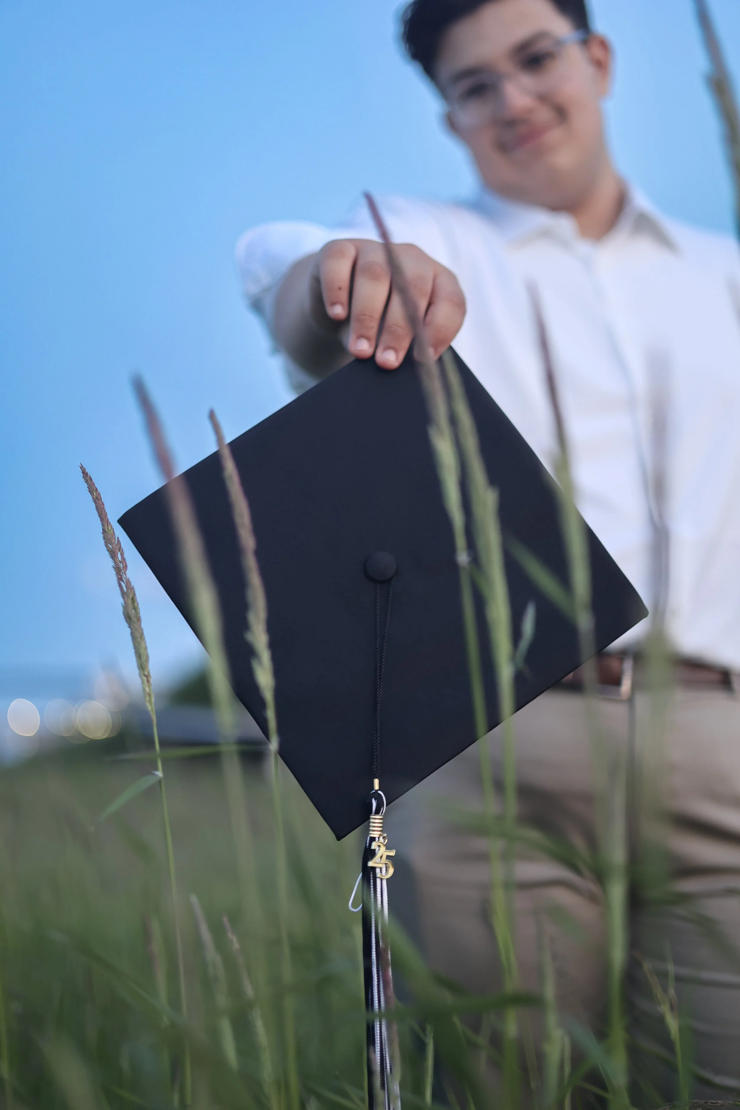 Young man wearing glasses and a white shirt holding a black graduation cap outdoors, with tall grass in foreground and blue sky in background.