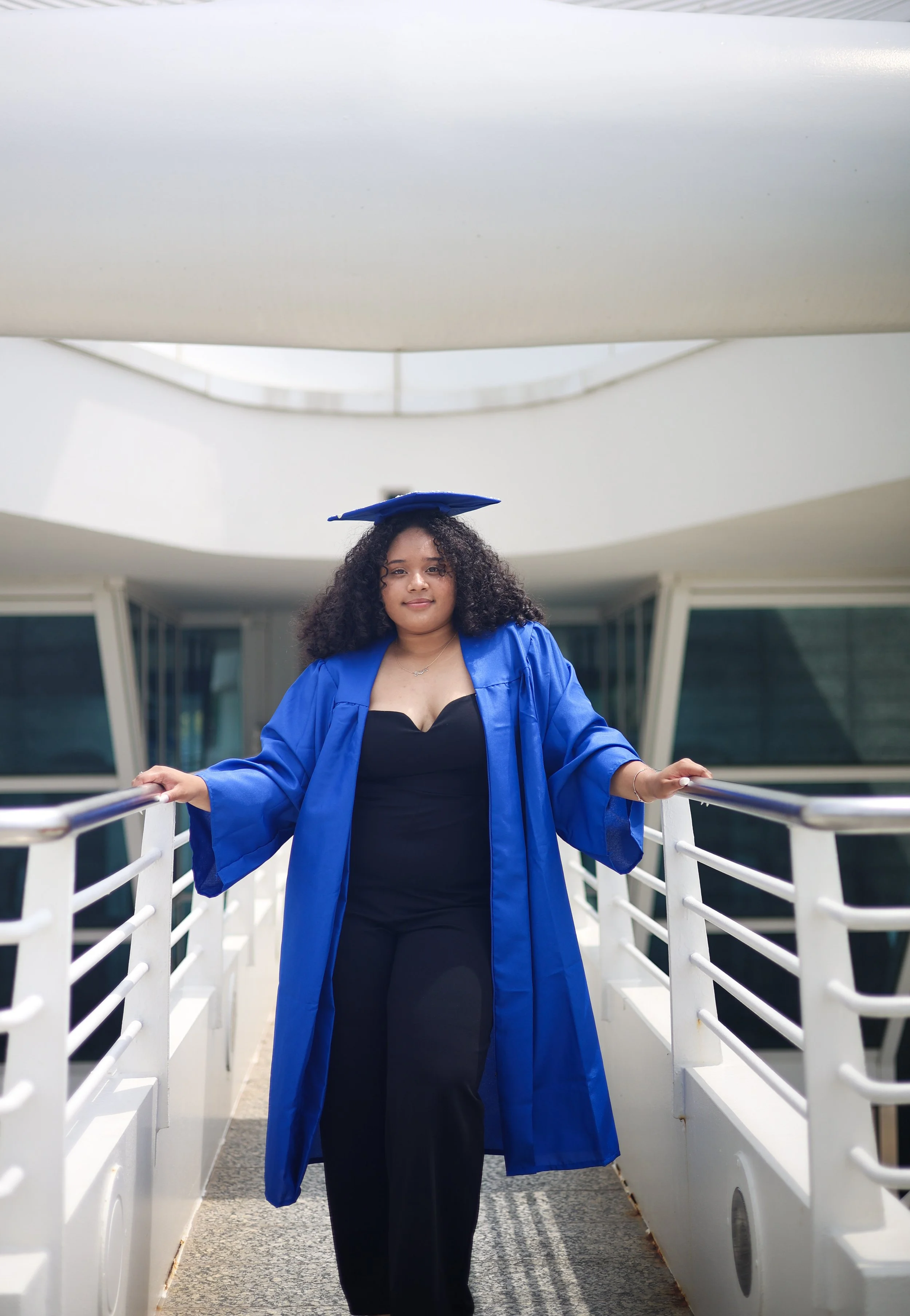 Young woman in blue graduation gown and cap standing on a bridge with her arms resting on the railing outside a modern building.