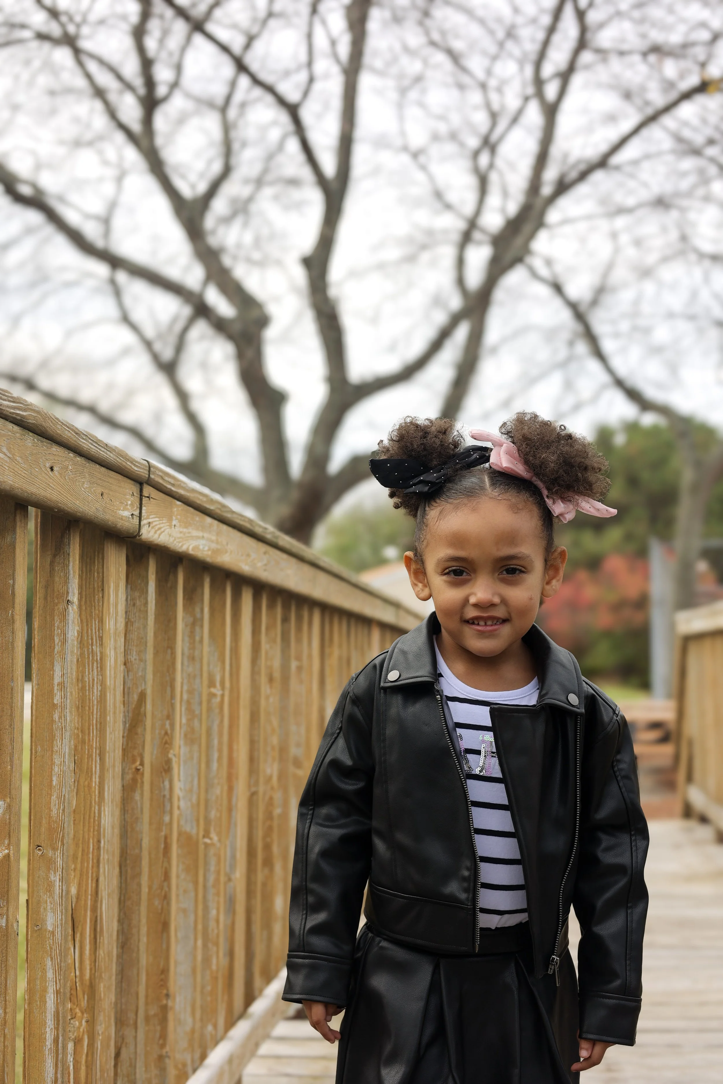 A young girl with curly hair tied with pink and black bows, wearing a black leather jacket over a black and white striped shirt, standing outside on a wooden walkway with a wooden fence and large bare tree in the background.