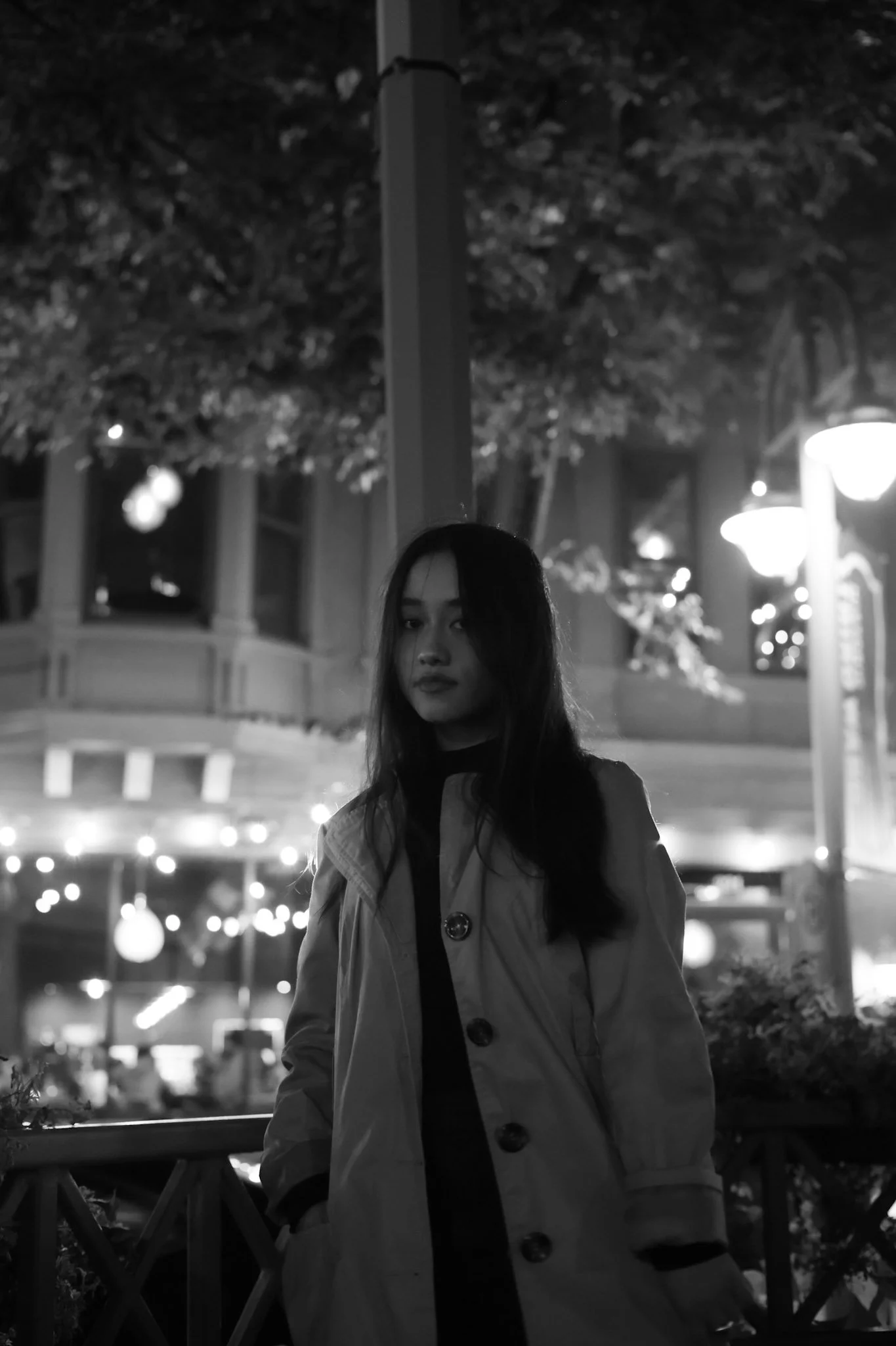 A young woman with long hair standing outside at night. She is wearing a coat and looking at the camera. The background has blurred street lights and trees.