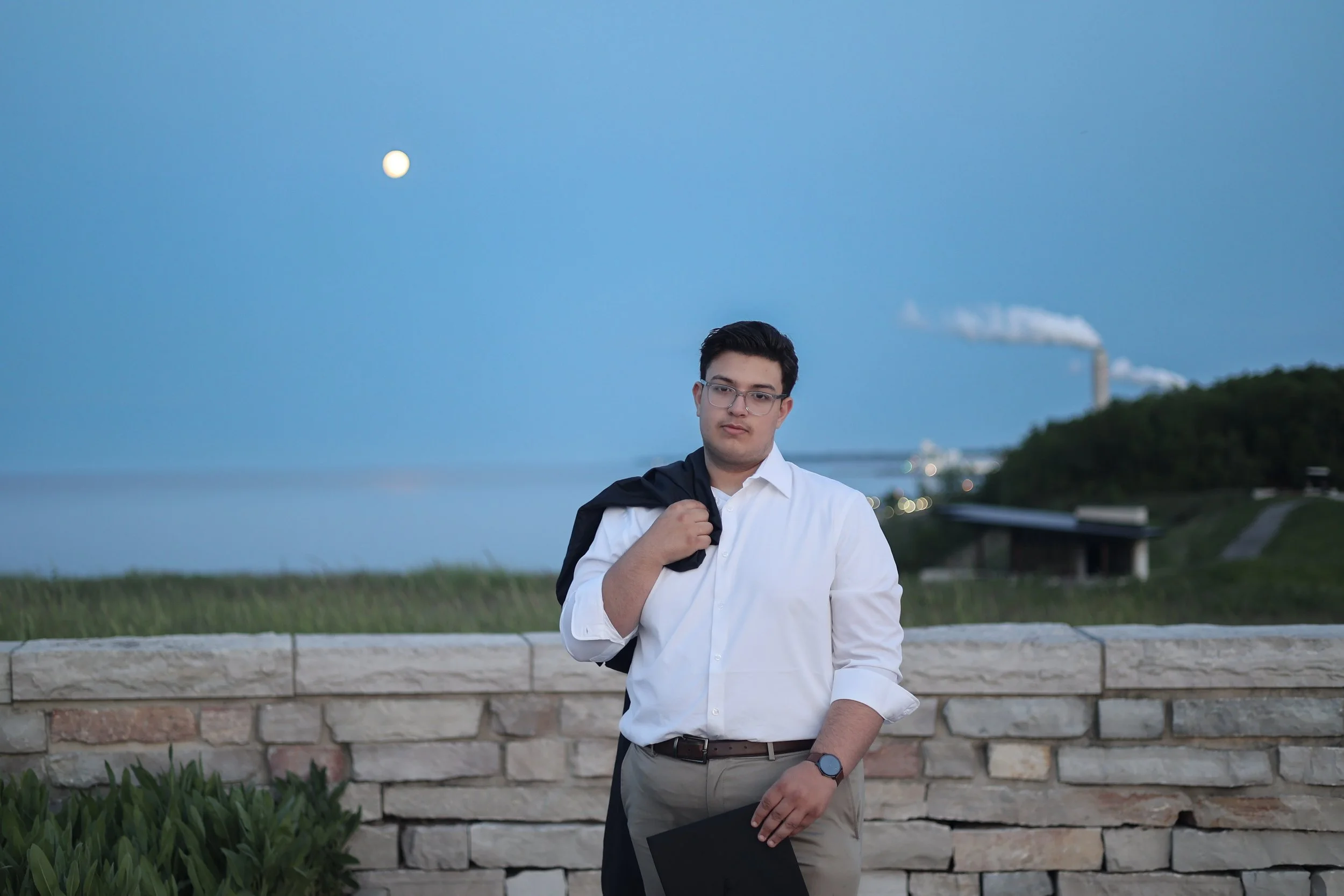 A young man with glasses in a white shirt and khaki pants standing outdoors by a stone wall, holding a black folder and a jacket over his shoulder, with a body of water and a full moon in the sky behind him.