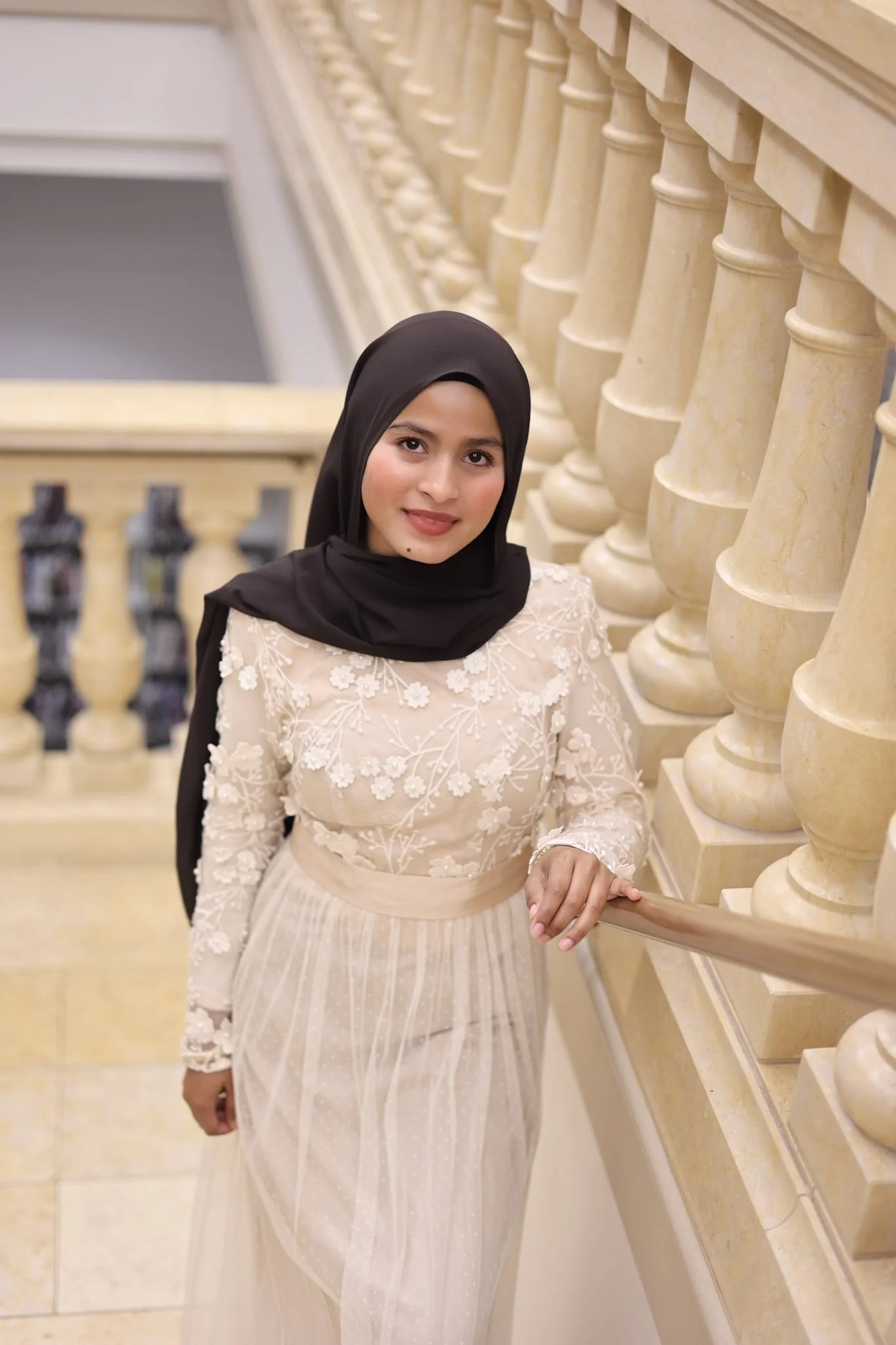 A woman in a beige dress with floral embroidery, wearing a black hijab, standing on a staircase with beige balusters, and she is holding the handrail.