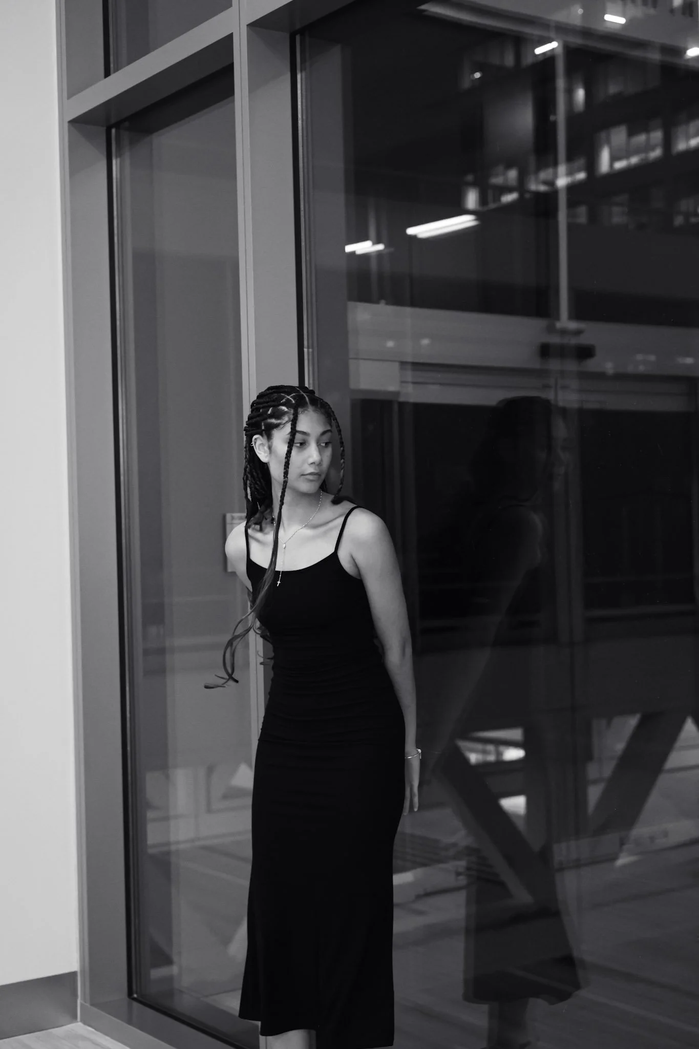 Young woman with braided hair in a black dress standing by a glass window, looking to the side.