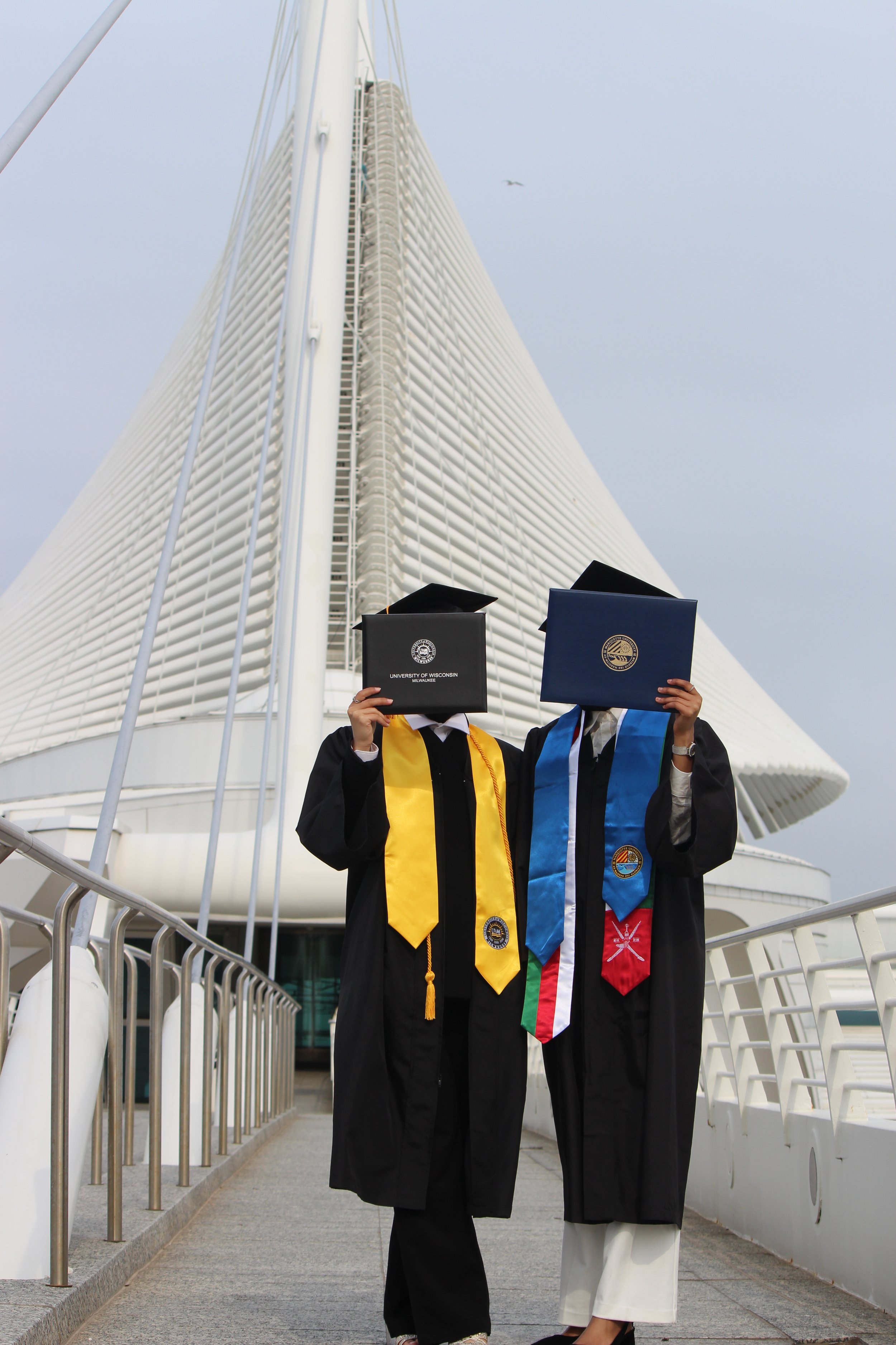 Two graduates in cap and gown holding diplomas, standing on a walkway in front of a modern architectural building, one wearing a yellow sash and the other with an international flag sash, both posing with the diplomas covering their faces.