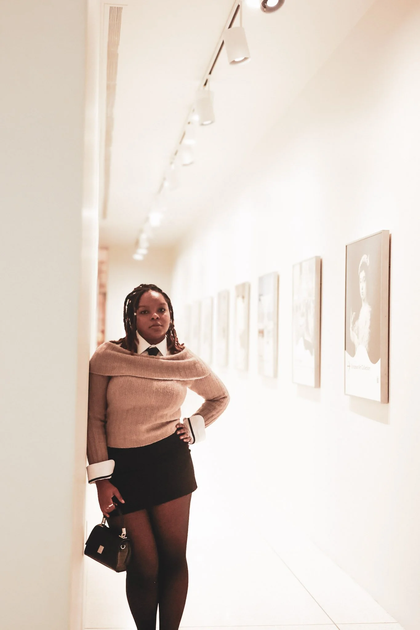 A woman with dark hair standing in an art gallery, posing with her hand on her hip. She is wearing a beige sweater, a black skirt, sheer tights, and holding a small handbag.