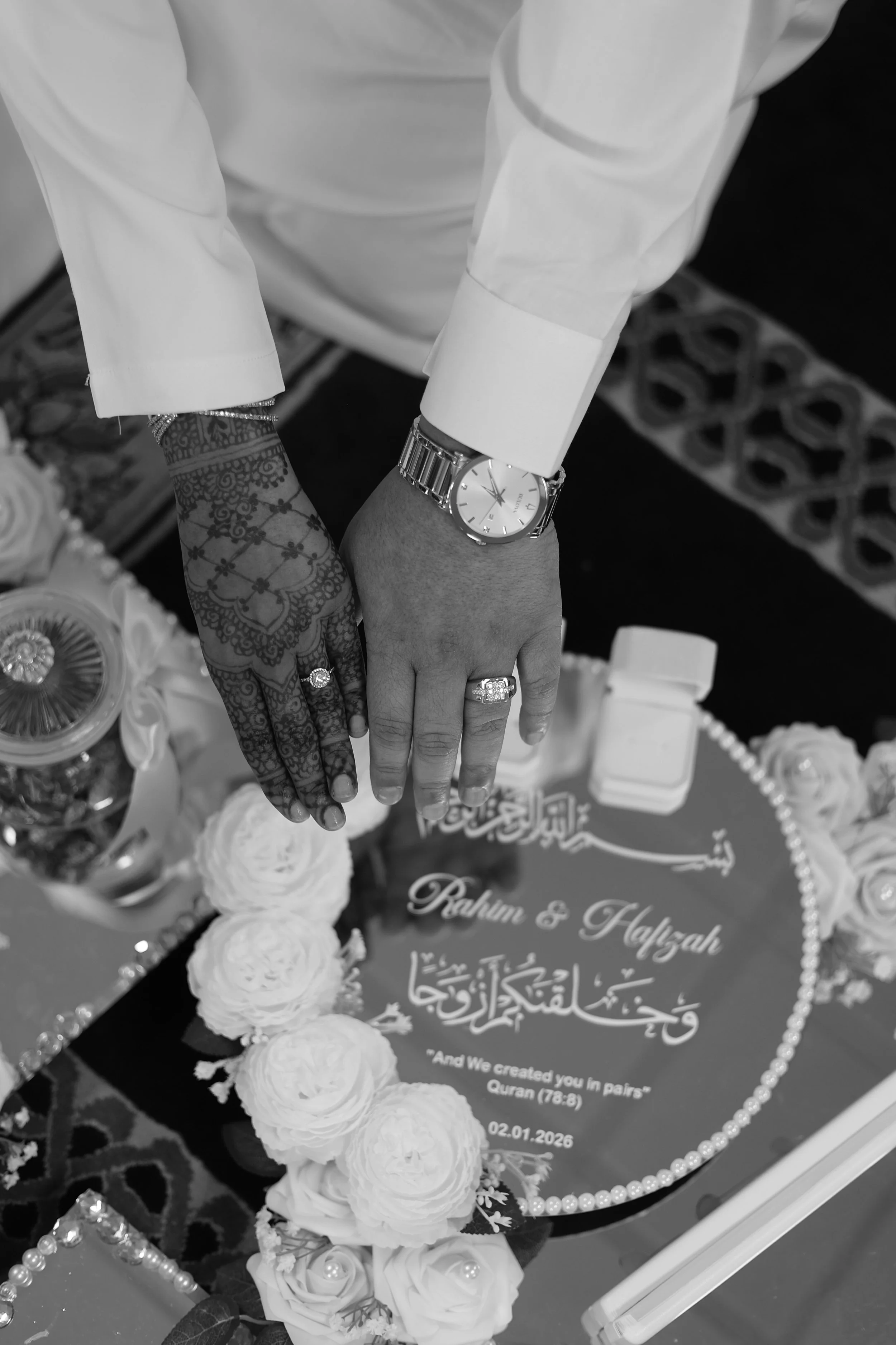 Close-up of two hands with wedding rings and a watch, touching over a decorated table with flowers and a card with Arabic script and English text about creating in pairs from the Quran, dated 02.01.2026.