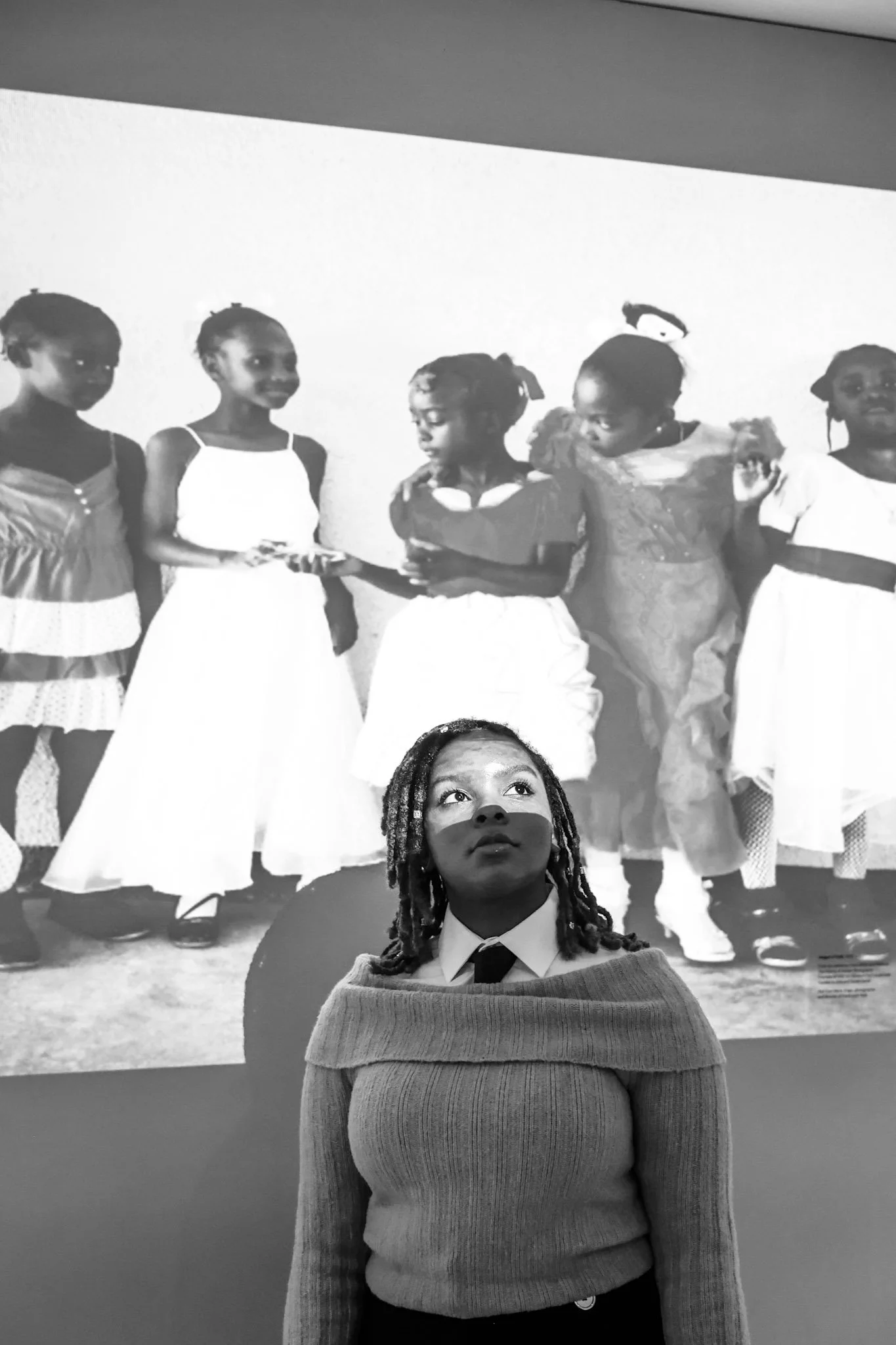Black woman with braided hair and a sweater poses in front of a large projection of a group of children on a white wall.