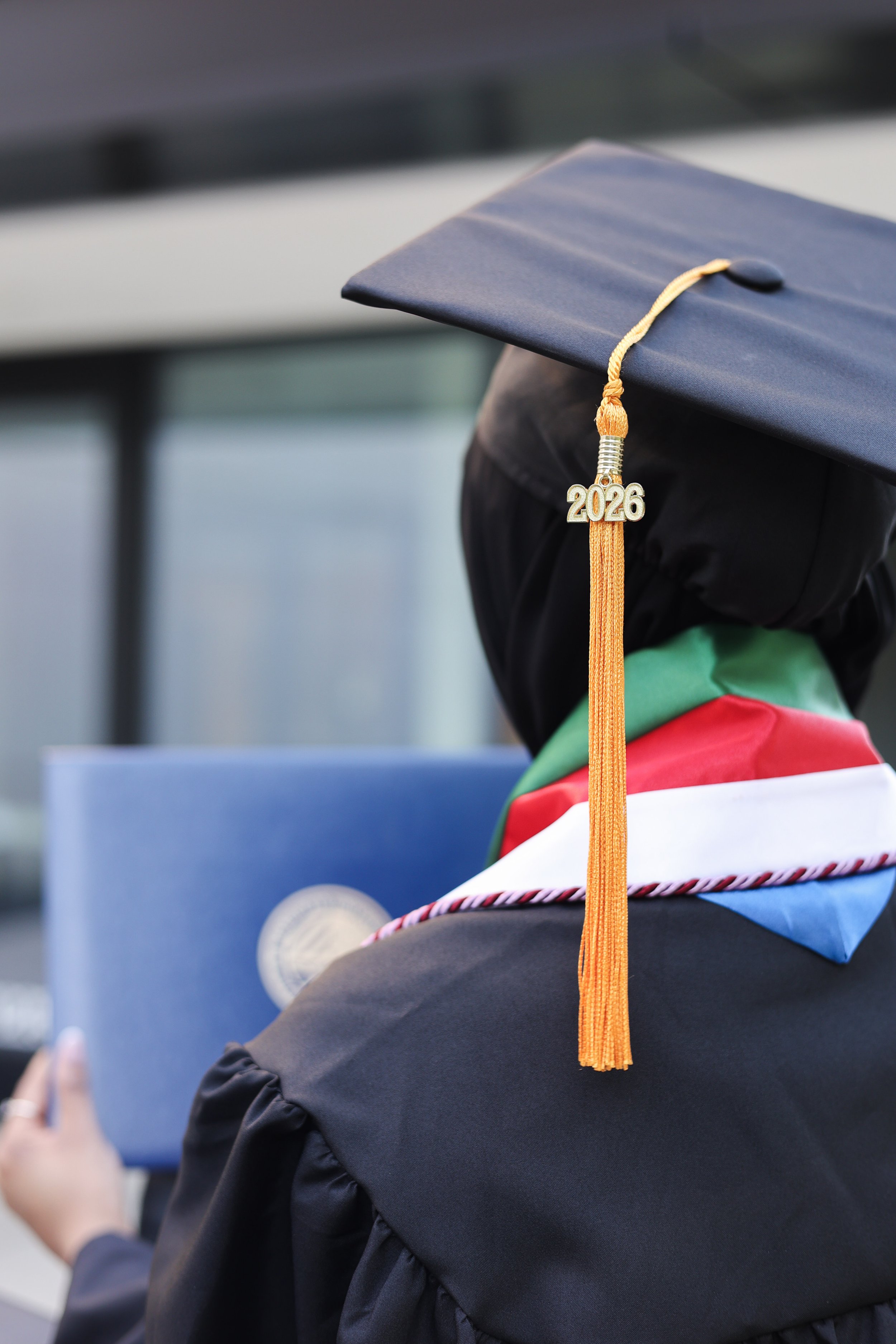 A graduation cap with a tassel displaying the year 2026, worn by a graduate dressed in a black gown and a colorful stoles, at a graduation ceremony.