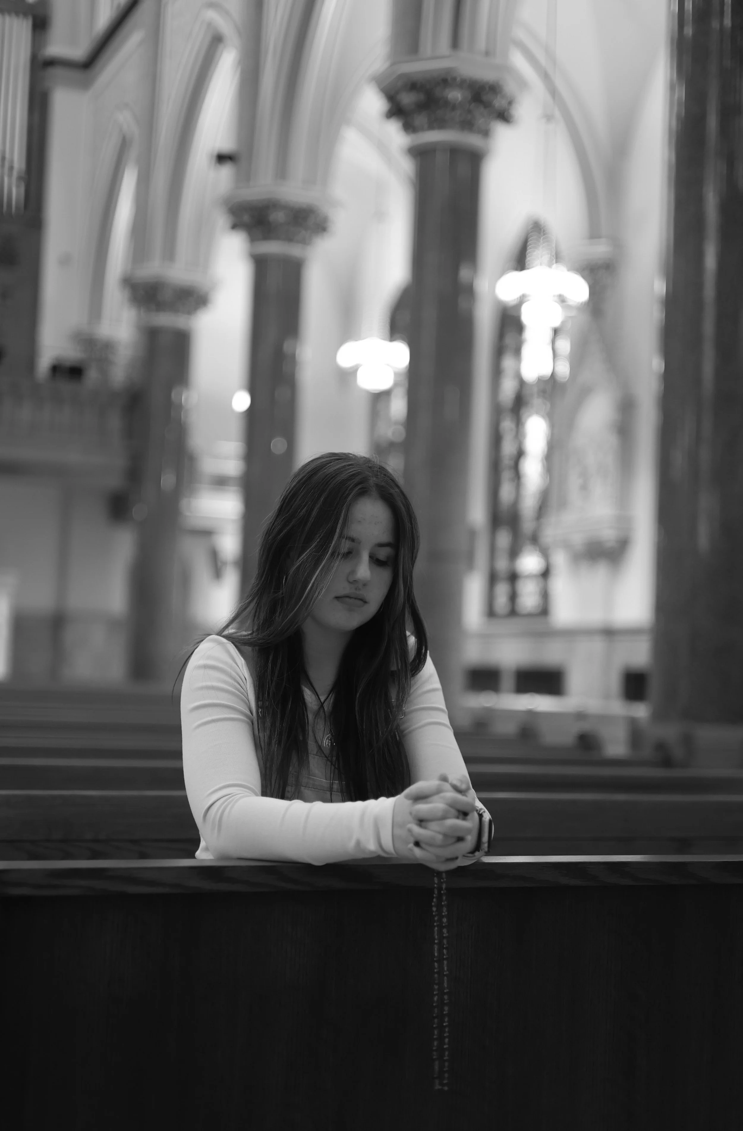 A young woman with long dark hair sitting at a church pew, hands clasped and holding a rosary, with a blurred background of church architecture.