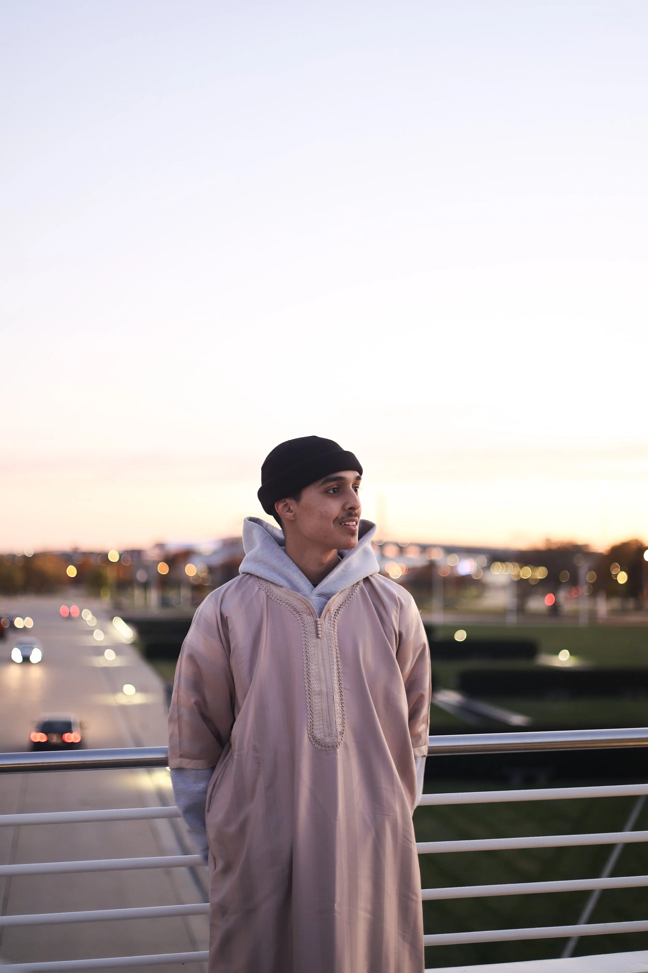 Young man wearing a beige traditional garment and black beanie standing on a bridge at sunset, with a cityscape and traffic blurred in the background.