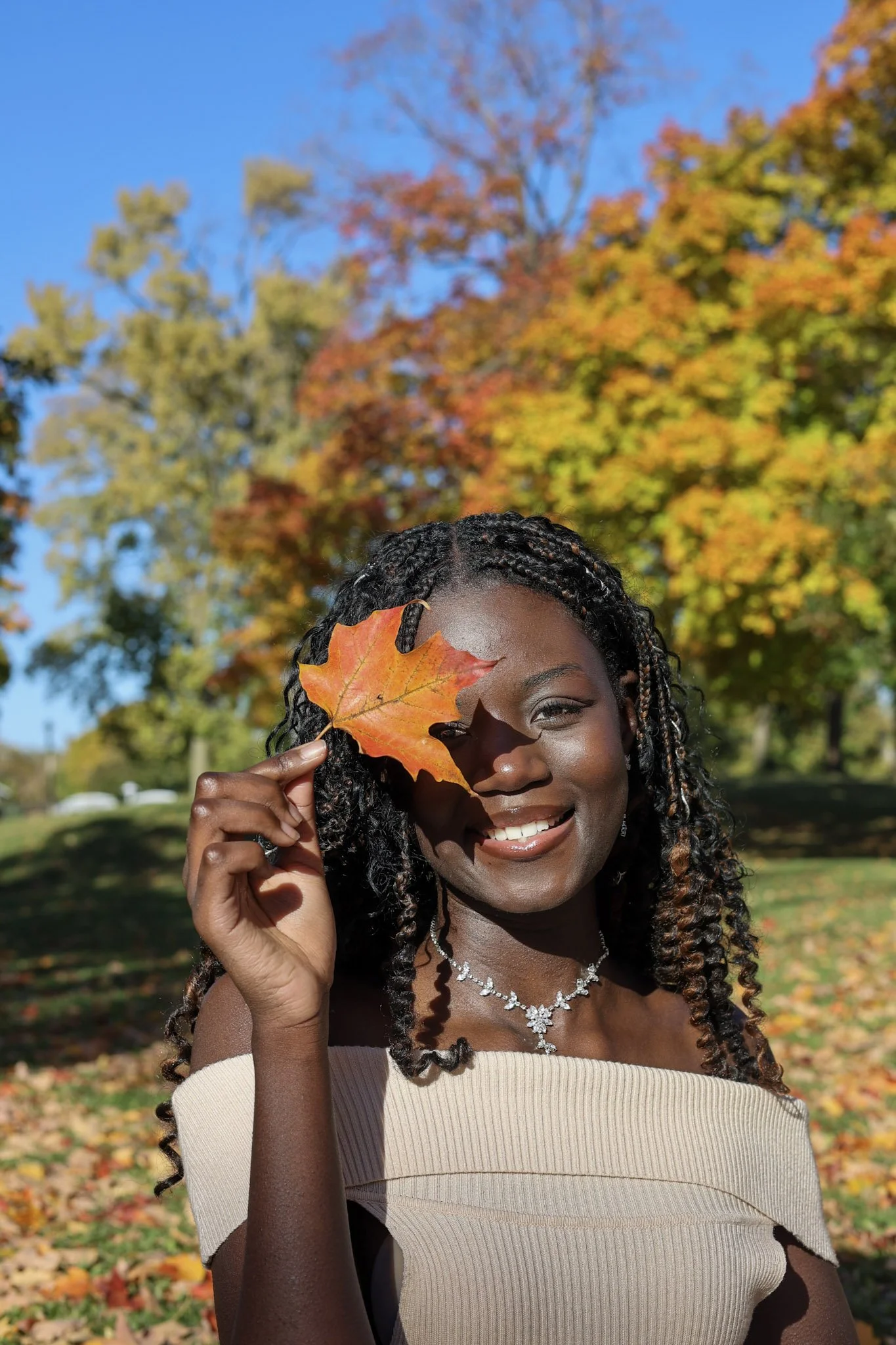 A smiling Black woman with braids, wearing a beige off-the-shoulder top and a silver necklace, holding an orange autumn leaf in front of her face, outdoors in a park with colorful fall foliage and clear blue sky in the background.