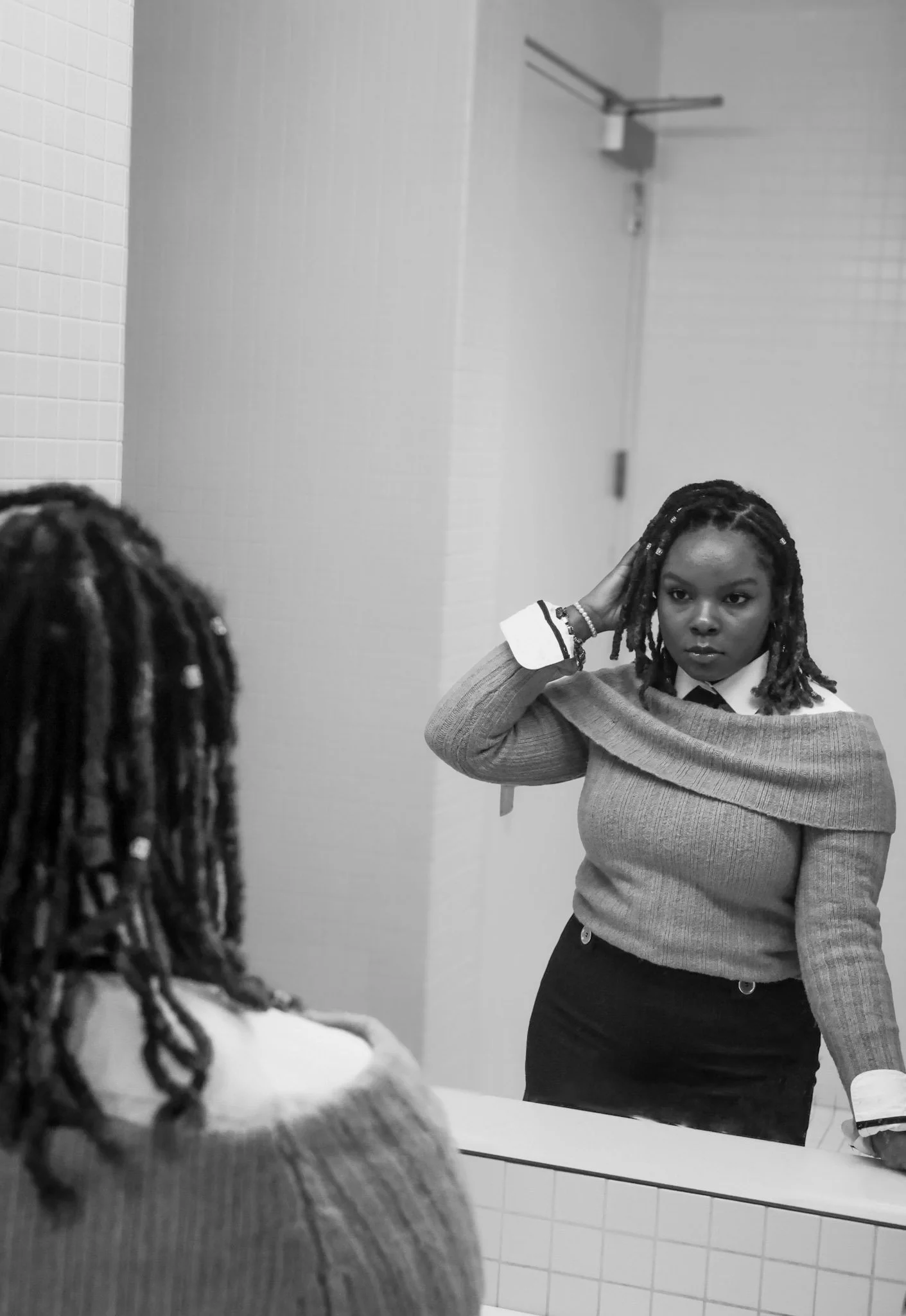 A woman with braided hair, wearing a gray sweater and black skirt, looking at herself in the mirror, adjusting her hair with a serious expression. She is seen in a bathroom with tiled walls.