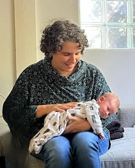 A woman with curly gray hair smiling while holding a newborn baby in her lap inside a house with a window in the background.