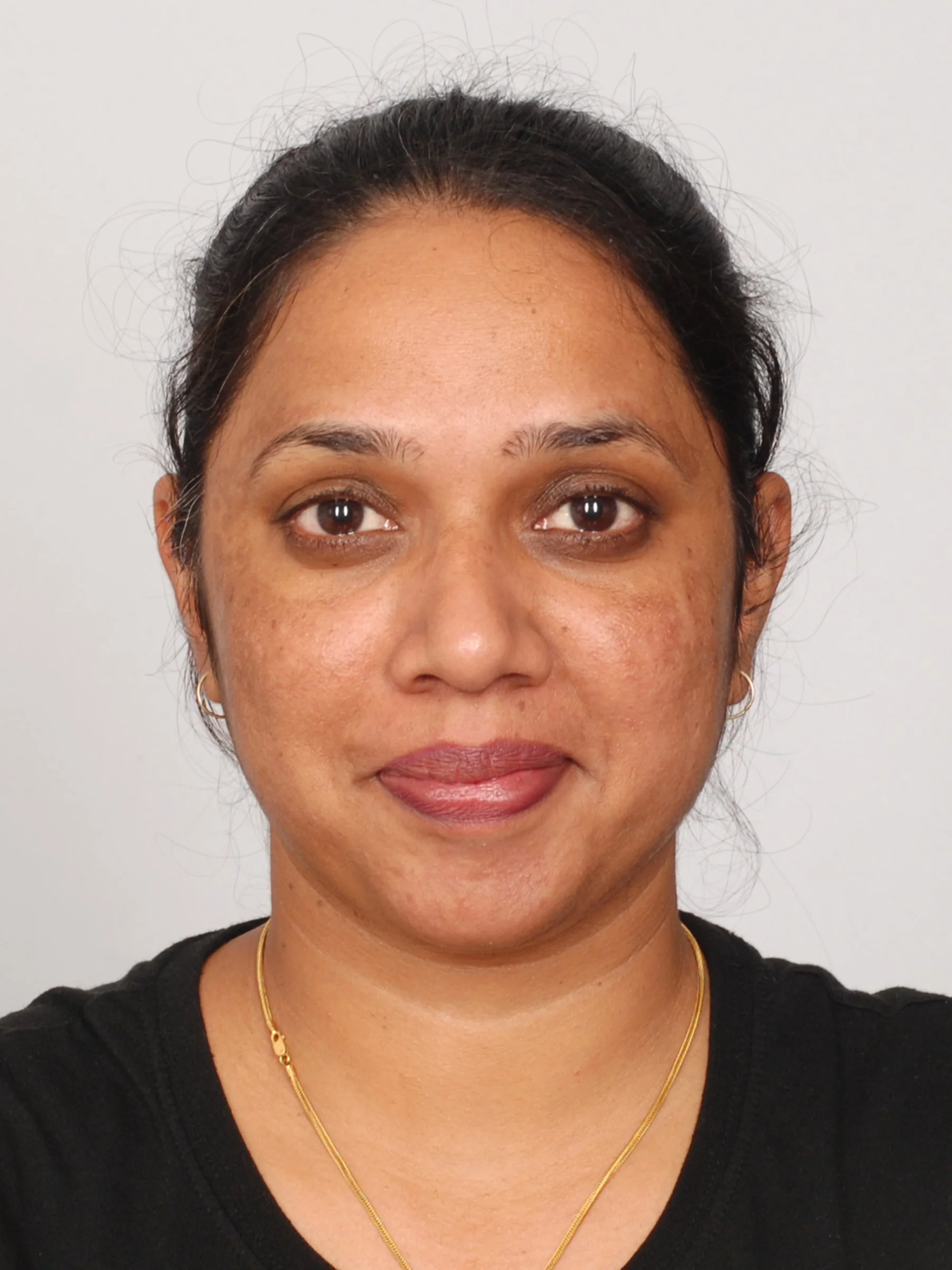 Front view of a woman with medium skin tone, dark hair pulled back, smiling, wearing a black shirt, gold necklace, and small hoop earrings, standing against a plain white background.