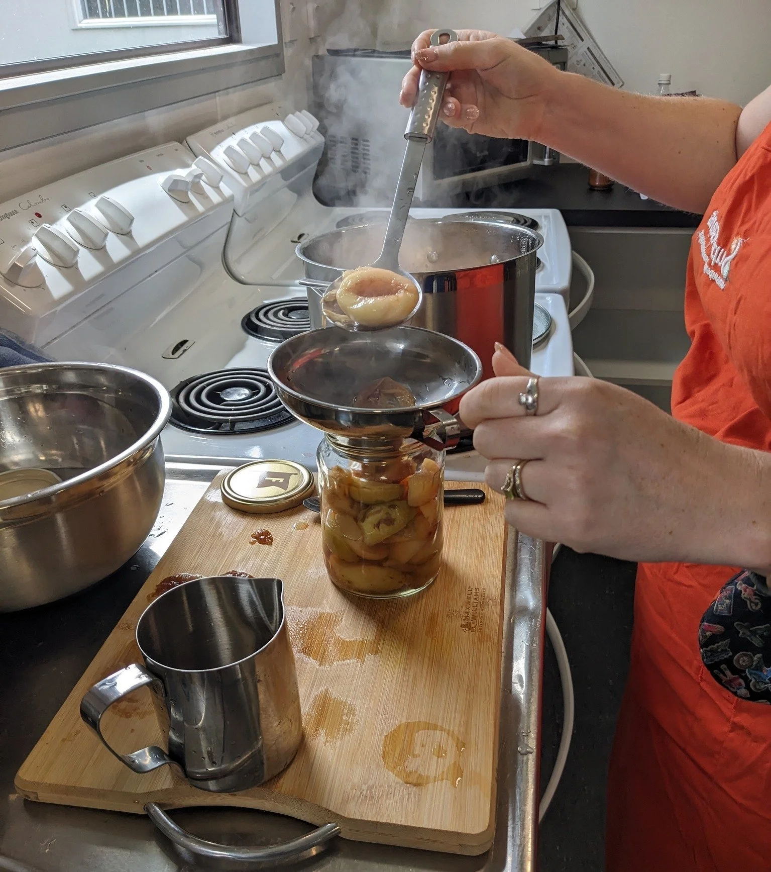 A person using a canning ladle to remove peaches from a jar filled with peach slices submerged in liquid, on a kitchen countertop. There are two metal measuring cups, a stainless steel mixing bowl, and a spice jar nearby. Steam rises from a boiling pot on the stove.