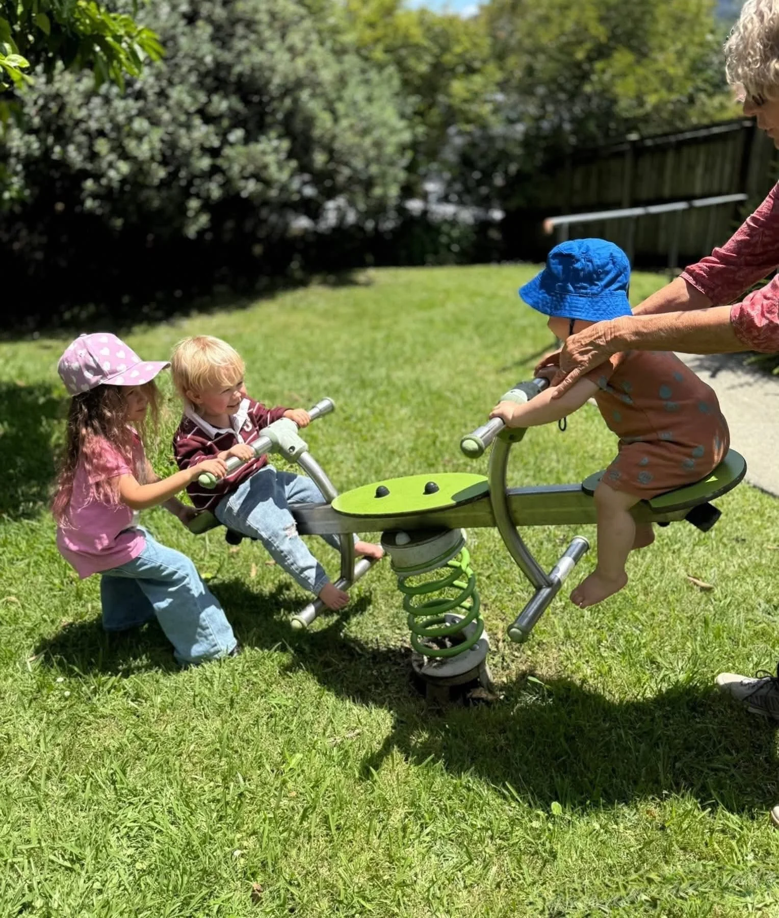 Three children playing on a spring rider in a grassy outdoor area, with an adult supervising. The children wear hats and casual clothing, and the scene is bright and sunny with trees and a fence in the background.