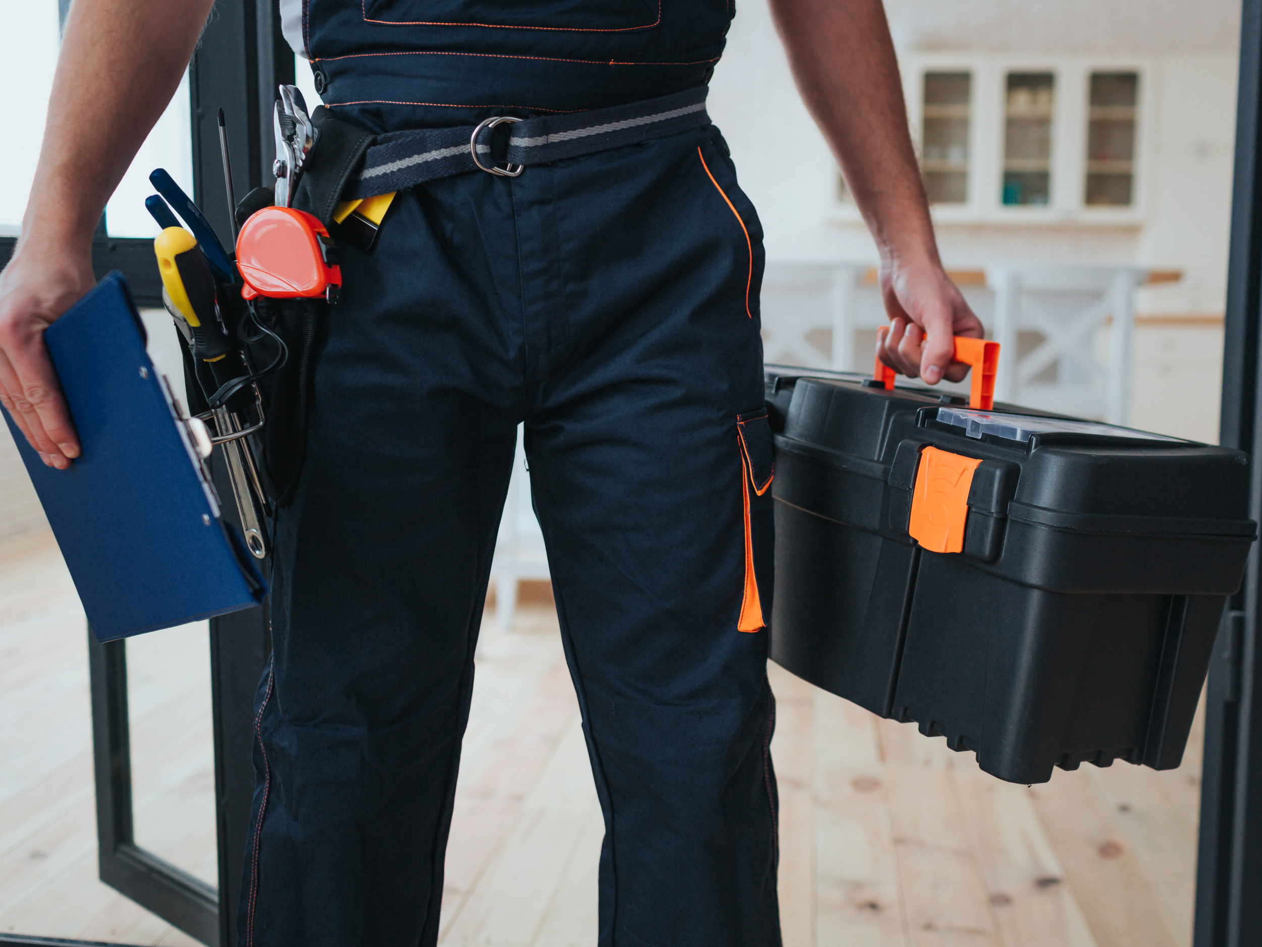 A person wearing dark work pants with orange stitching holding a black toolbox with a yellow handle and a leg pouch with tools attached, including a tape measure, pliers, and a screwdriver.